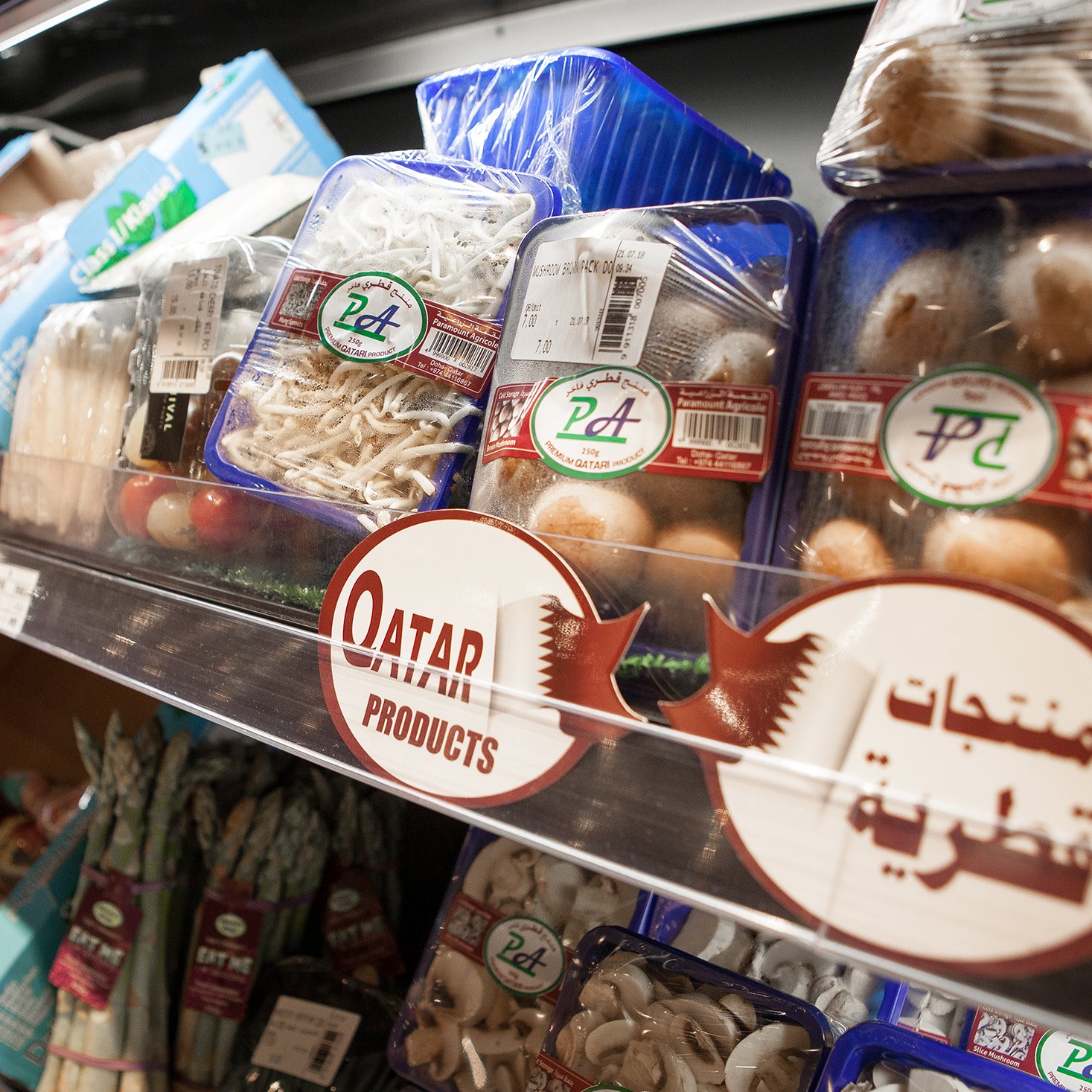 L'image montre une étagère de produits alimentaires dans un supermarché. On peut voir des emballages transparents contenant différentes sortes de champignons et d'autres légumes. L'étiquette "Qatar Products" est également présente, indiquant que ces produits proviennent du Qatar. On aperçoit des sachets d'autres légumes, notamment des asperges et des algues, organisés de manière ordonnée sur l'étagère. L'éclairage est clair, mettant en valeur la fraîcheur des produits.
