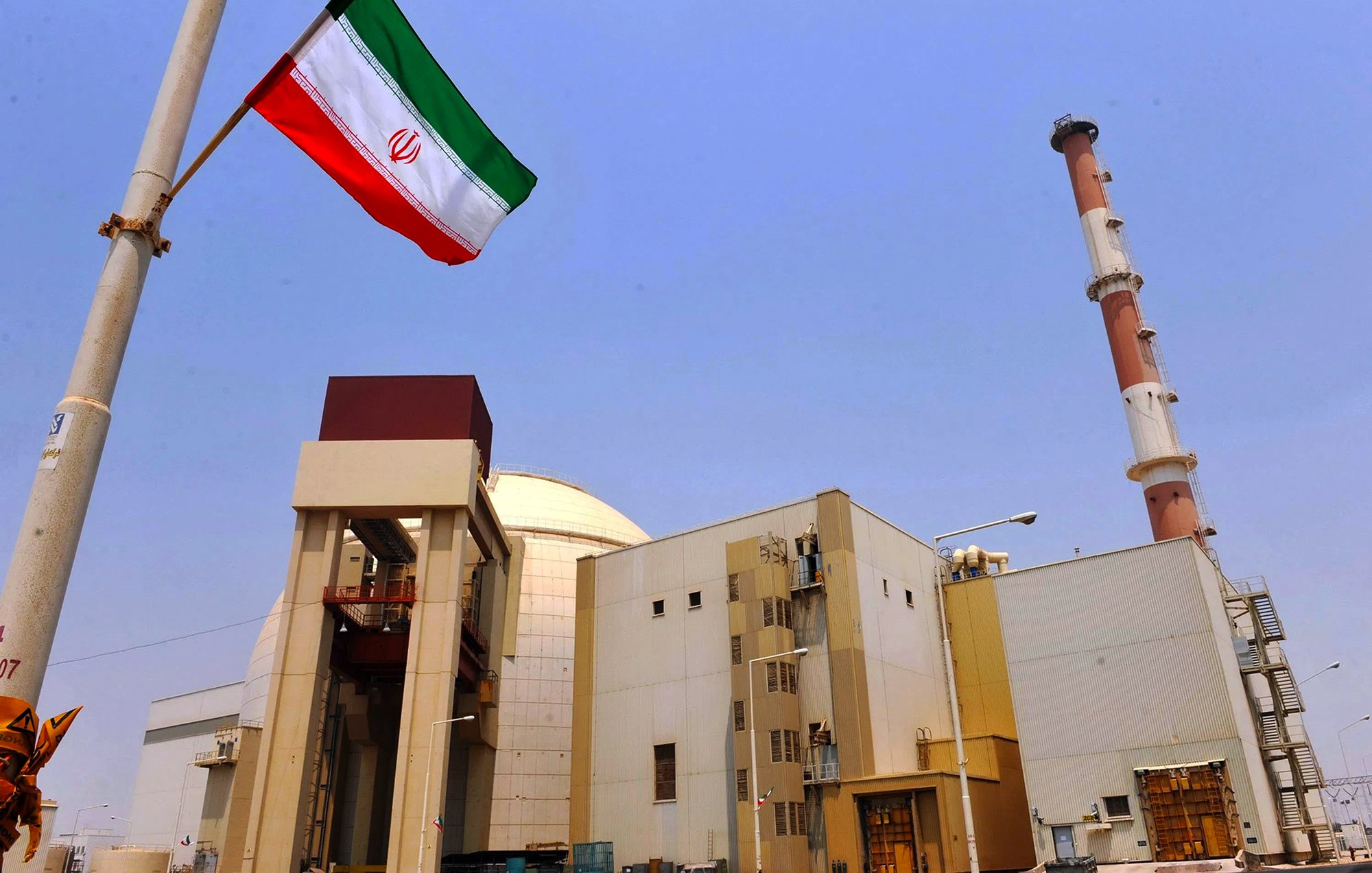 L'image montre une installation industrielle avec des structures modernes. On peut voir une grande cheminée au fond et un bâtiment principal qui semble être lié à l'énergie. Au premier plan, il y a un mât avec le drapeau iranien flottant, qui arbore des bandes rouges, blanches et vertes. Le ciel est dégagé, créant un contraste avec les bâtiments. L'ensemble évoque une infrastructure liée à la production d'énergie, probablement nucléaire.