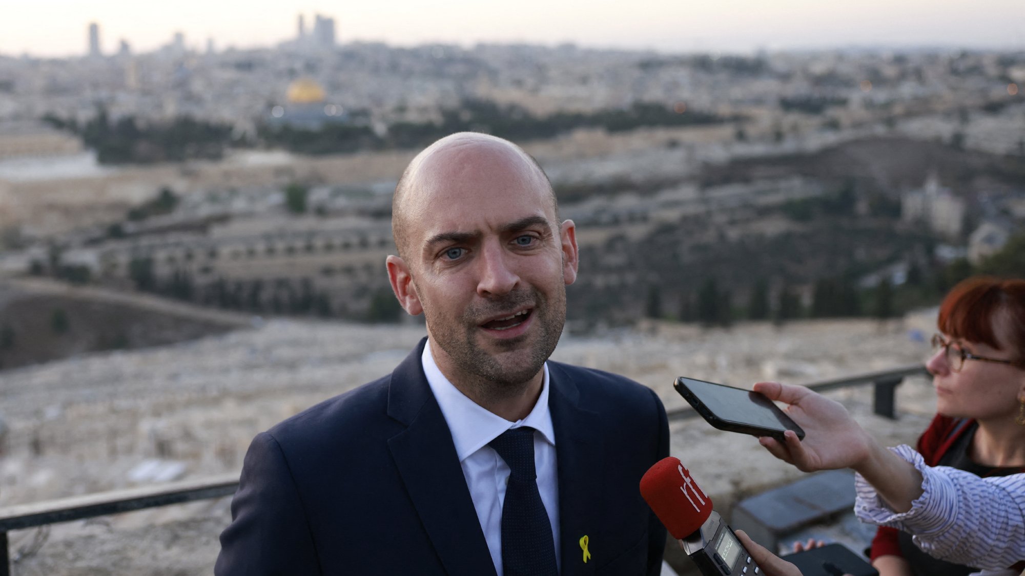 L'image montre un homme en costume, probablement un porte-parole ou un responsable politique, s'exprimant devant un groupe de journalistes. Il se tient à l'extérieur avec une vue panoramique de la ville en arrière-plan, qui semble être Jérusalem, avec des bâtiments et un paysage urbain. Le ciel est clair, et la lumière du crépuscule donne une atmosphère calme à la scène. Plusieurs microphones et téléphones montrent l'intérêt médiatique pour ses déclarations.