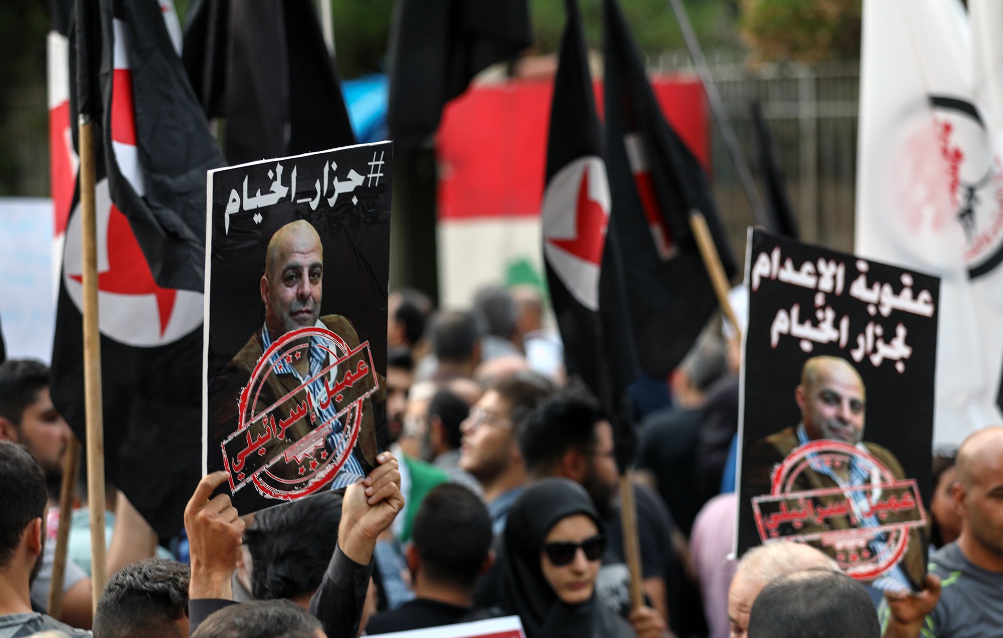 The image depicts a crowd of protesters holding signs and flags. The signs feature a portrait of a man along with a hashtag and phrases in Arabic, indicating a political or social issue. The crowd appears engaged and is likely part of a demonstration. The flags are predominantly black with red symbols, contributing to the atmosphere of the protest. The environment suggests a public gathering, possibly in response to a recent event or issue.