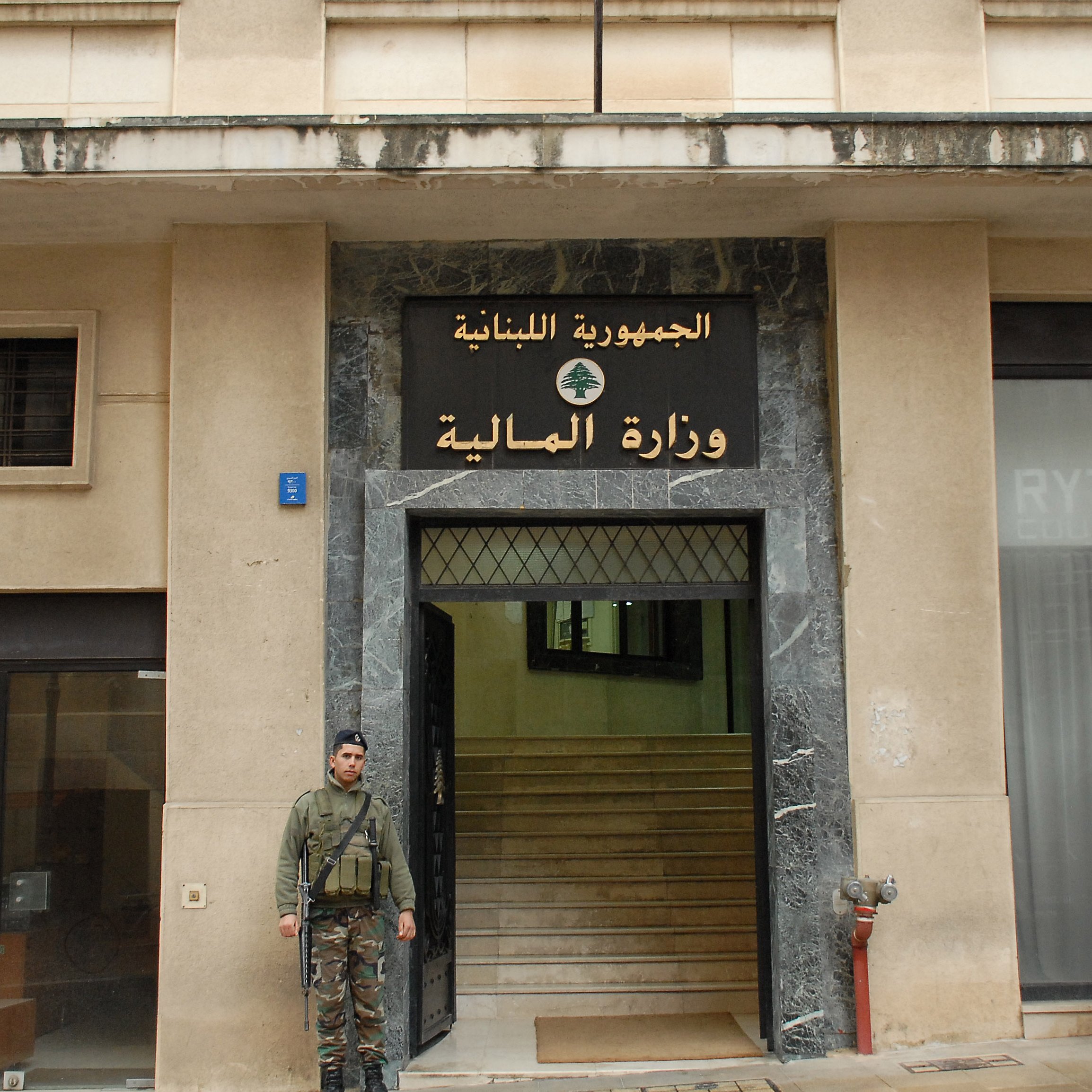 The image shows a soldier standing outside a building with a large sign displaying the words "الجمهورية اللبنانية" (Lebanese Republic) and "وزارة المالية" (Ministry of Finance) in Arabic. The building has a modern façade with a marble entrance. The soldier is wearing military uniform and holding a weapon, indicating a security presence at the site. The overall setting suggests a government location in Lebanon.