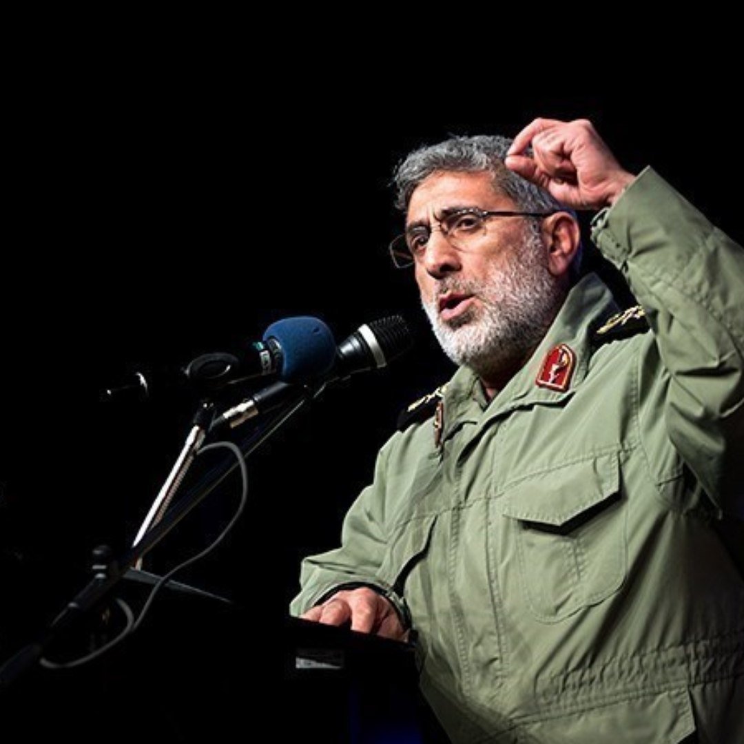 L'image montre un homme en uniforme militaire, debout devant un micro sur une scène. Il semble s'adresser à un public, probablement en train de faire un discours. Son expression est sérieuse, et il gesticule avec la main pour soutenir ses propos. Sur le podium, on peut voir des fleurs ou des plantes qui ajoutent une touche décorative à la scène.