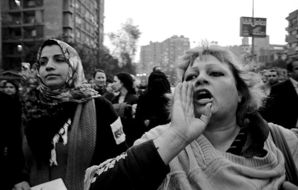 L'image montre une scène de manifestation en noir et blanc. Au premier plan, une femme au cheveux blonds, visiblement passionnée, crie ou exprime une émotion forte, tandis qu'une autre femme, à gauche, porte un foulard et semble attentive. On aperçoit en arrière-plan un grand groupe de personnes, ce qui suggère un mouvement collectif. L'atmosphère est chargée d'énergie et de détermination. Les bâtiments autour ajoutent un contexte urbain à cette démonstration.