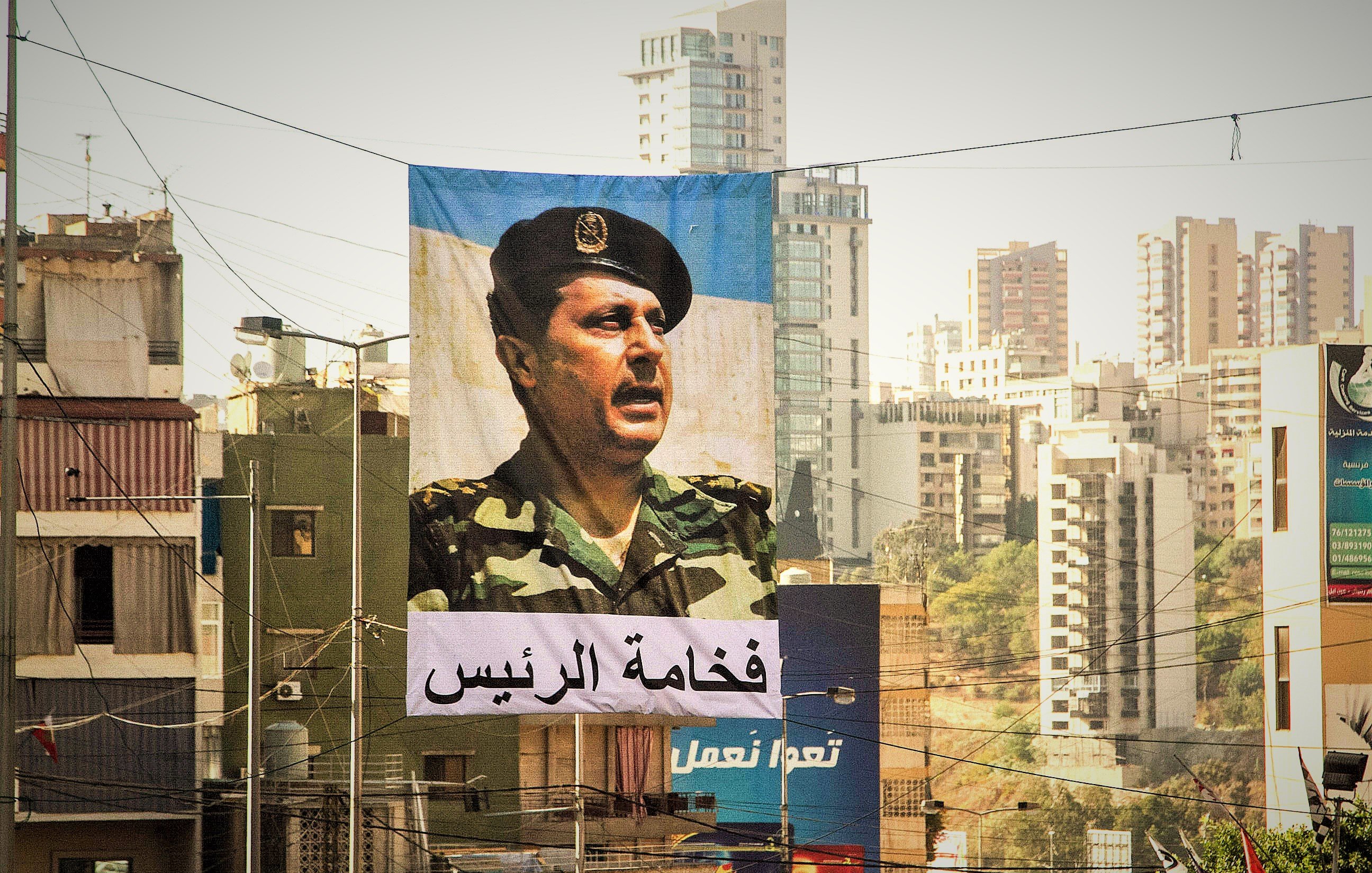 L'image montre une grande bannière suspendue dans une rue urbaine. Sur cette bannière, un portrait d'un homme en uniforme militaire est visible, face à un public. En dessous de l'image, il y a un texte en arabe qui semble faire référence à une autorité ou un leadership. En arrière-plan, on aperçoit des bâtiments modernes et d'autres structures urbaines typiques d'une ville. La lumière est douce, ce qui donne une atmosphère particulière à la scène.