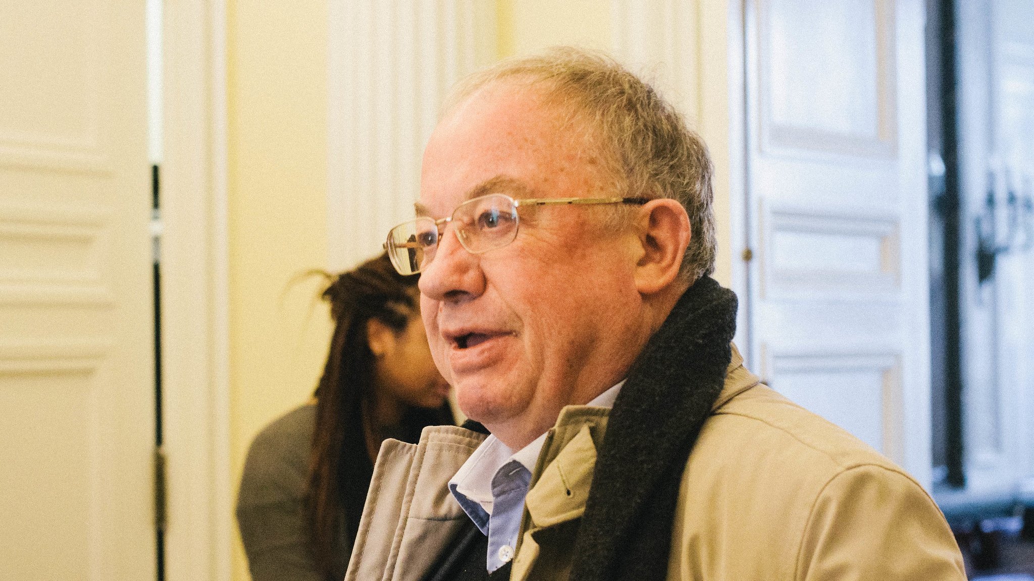 Homme âgé avec des lunettes, portant un manteau, dans un intérieur lumineux.