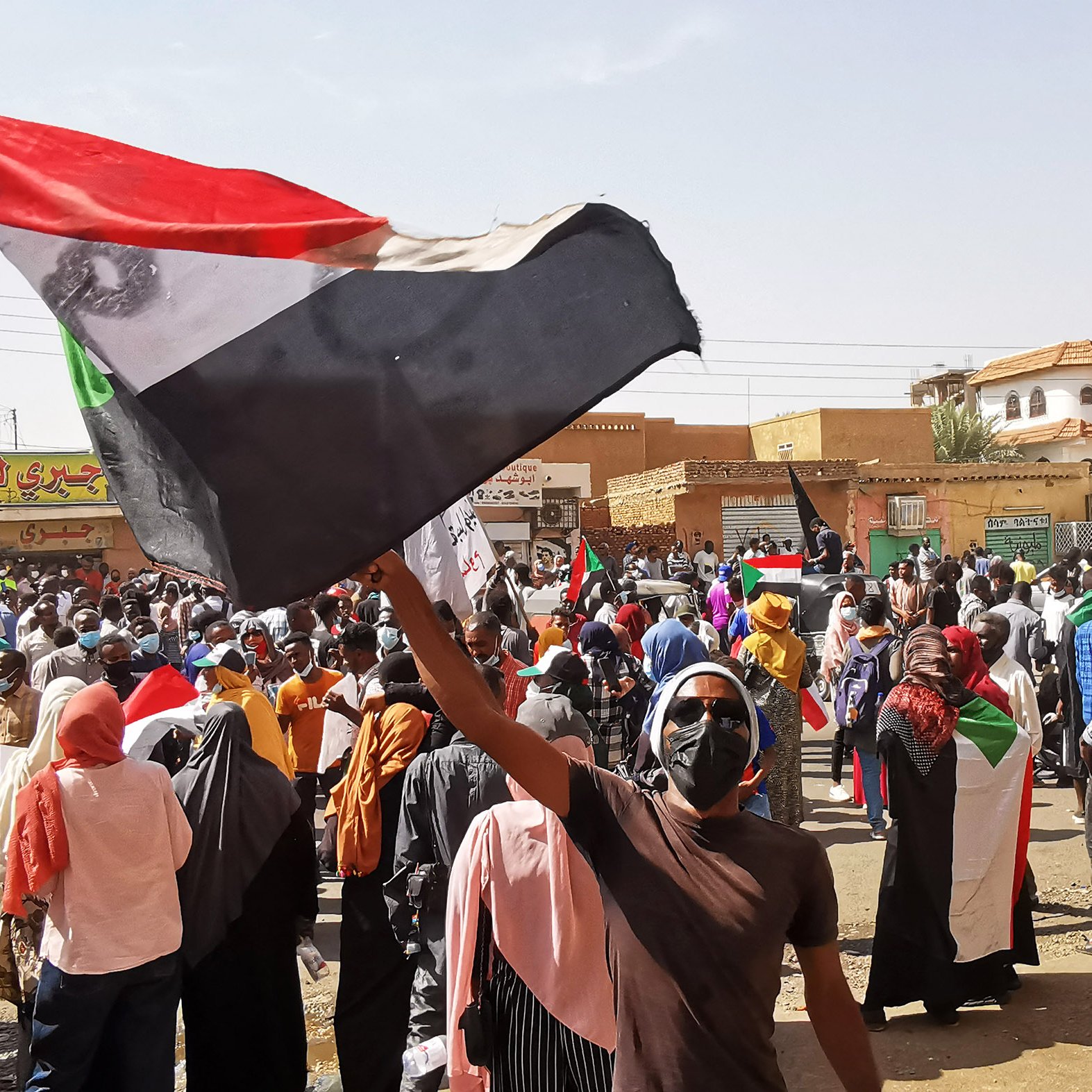 L'image montre une grande foule réunie dans une manifestation, avec des personnes brandissant des drapeaux. Au premier plan, un individu tient un drapeau soudanais, et de nombreuses autres personnes portent des masques. L'atmosphère semble énergique et mobilisée, avec des pancartes et des expressions de détermination. En arrière-plan, on peut apercevoir des bâtiments et des enseignes, indiquant un cadre urbain.
