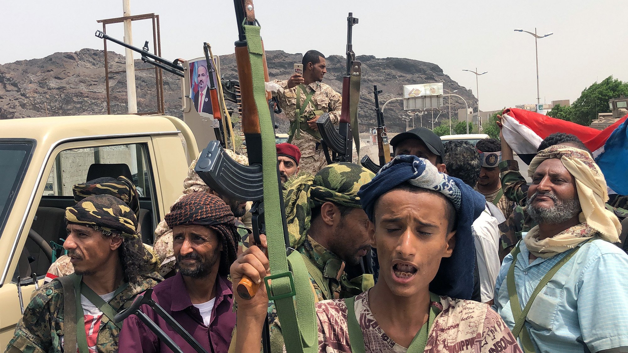 The image depicts a group of armed individuals standing together in an outdoor setting. They are dressed in military-style clothing and some wear traditional headscarves. Several of them are holding rifles and appear to be engaged in a rally or demonstration. The background features a vehicle and a large rock formation, with a hint of a cityscape. A flag is visible, adding to the atmosphere of the gathering, which seems to pertain to a political or military context. The expressions and postures of the individuals reflect a sense of determination or unity.