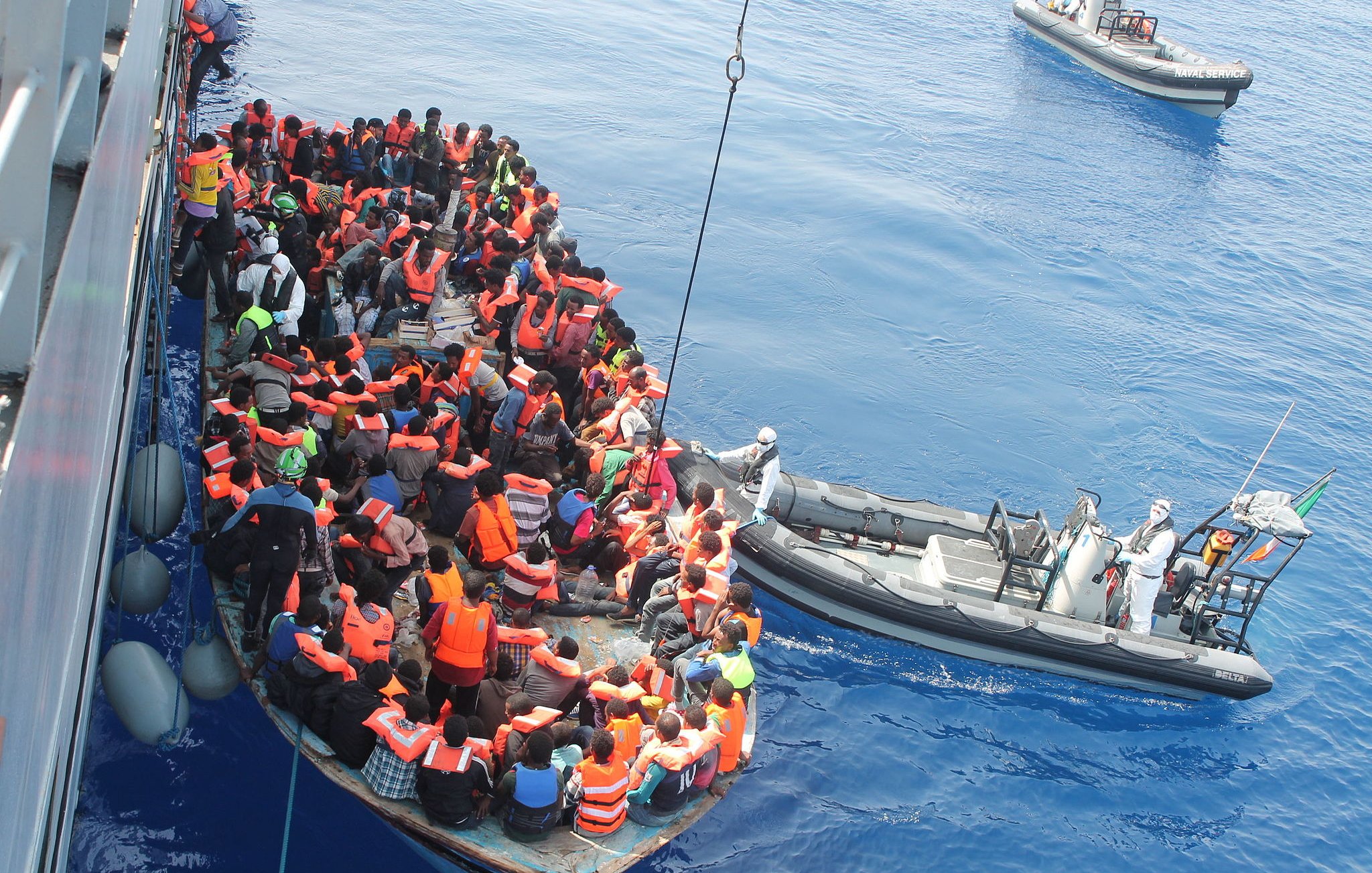 L'image montre un groupe de personnes dans une embarcation sur une mer calme. La plupart des individus portent des gilets de sauvetage orange, indiquant qu'ils sont en situation de danger. À proximité, une autre petite embarcation semble aider le groupe, probablement une équipe de secours. L'atmosphère générale évoque des enjeux humanitaires, liés à la migration et aux sauvetages en mer. Le ciel est clair et l'eau est d'un bleu profond.
