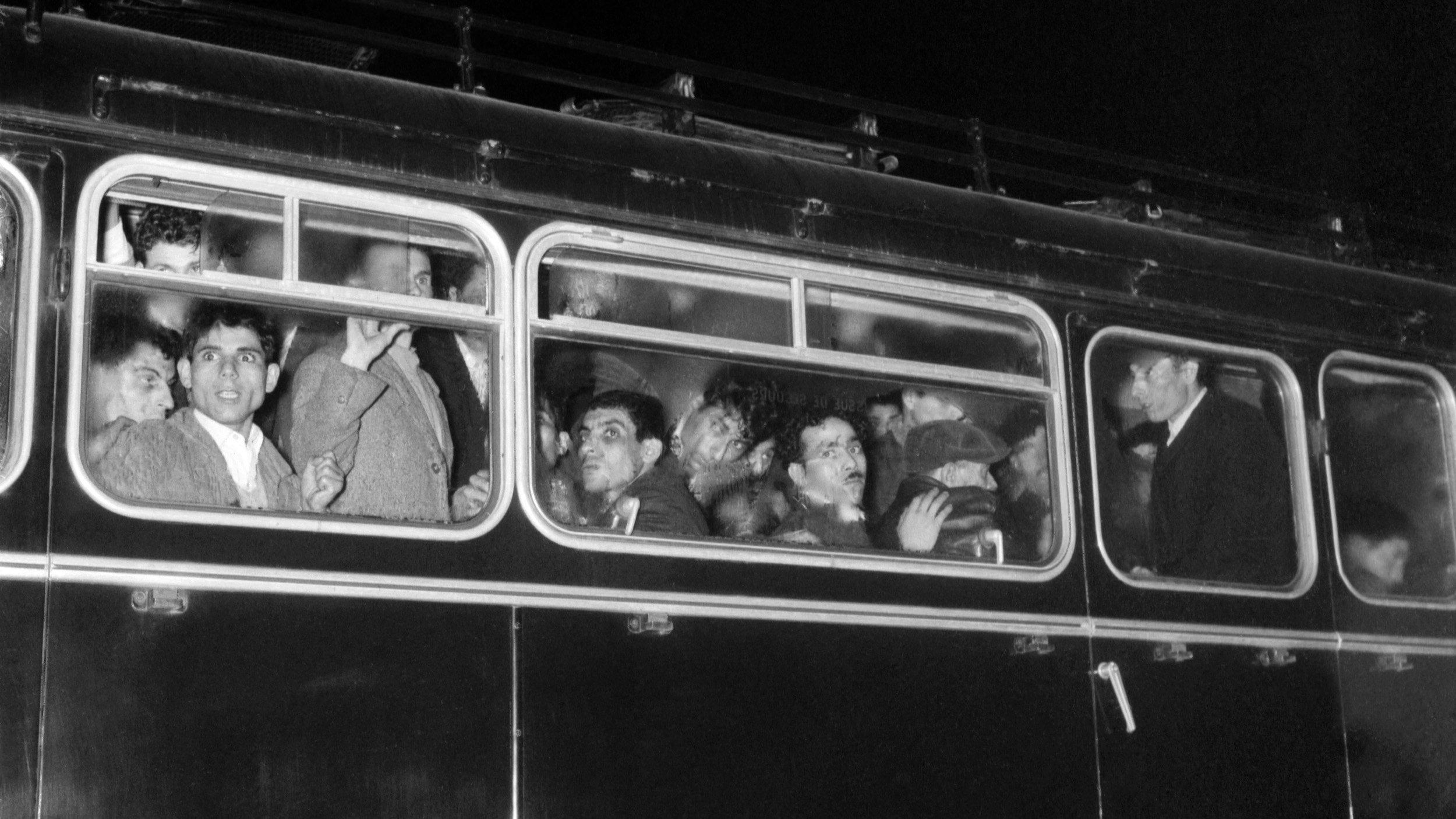 تصویر نشان‌دهنده یک اتوبوس است که در شب در حال حرکت است. در داخل اتوبوس، چندین نفر وجود دارند که از پنجره‌ها به بیرون نگاه می‌کنند. چهره‌های آن‌ها به وضوح قابل مشاهده است و به نظر می‌رسد در حال گفتگو یا ابراز احساساتند. طراحی اتوبوس و فضای اطراف، حالتی تاریخی و نوستالژیک دارد. رنگ تصویر به سیاه و سفید است که بر جذابیت آن افزوده و حس زمان گذشته را القا می‌کند.