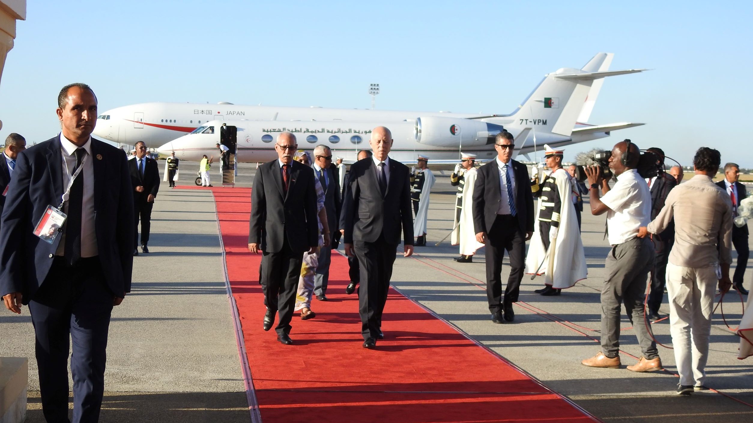 L'image montre un aéroport avec une piste. Un avion, visible en arrière-plan, est stationné. Des personnes marchent sur un tapis rouge, probablement des dignitaires ou des personnalités importantes. Certains hommes en costume accompagnent les personnes sur le tapis, tandis que d'autres, habillés en tenue formelle, semblent filmer ou prendre des photos de l'événement. L'atmosphère est formelle et festive, indiquant une arrivée importante.