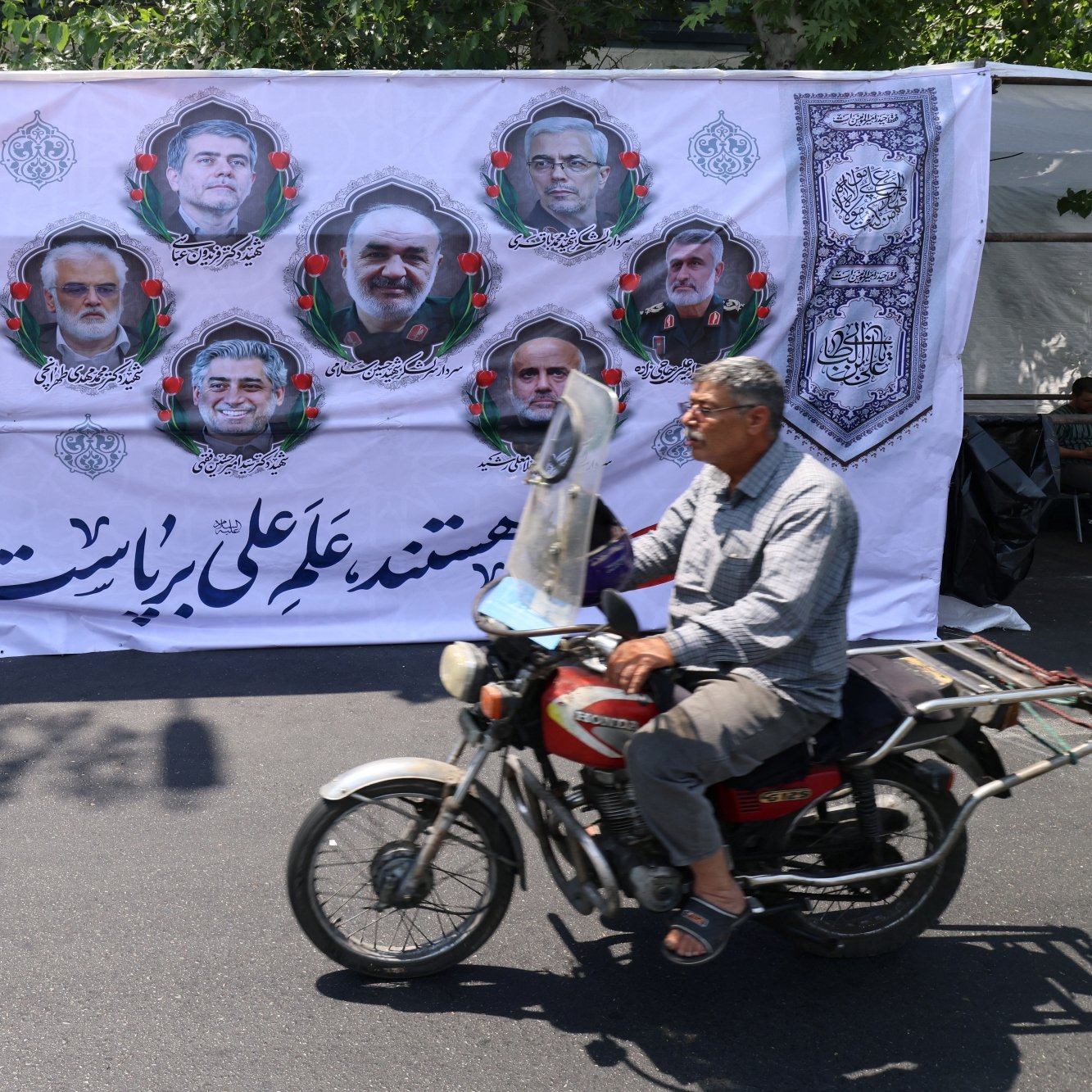 Un homme à moto passe devant une grande affiche avec des portraits et des motifs décoratifs.