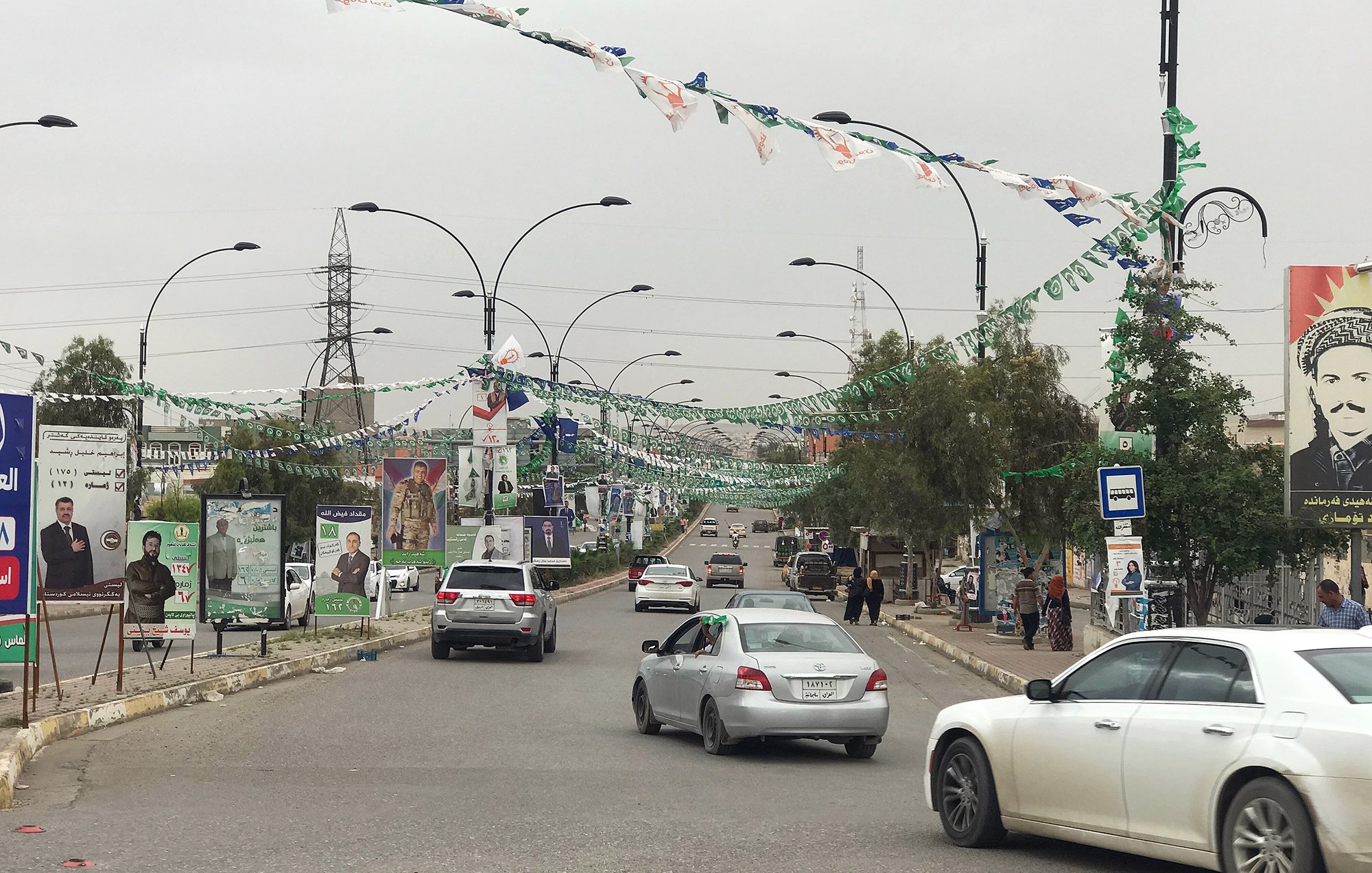 تظهر الصورة مشهداً لشارع في منطقة حضرية، حيث تنتشر الأعلام والزينة على أعمدة الإنارة. يمكن رؤية عدد من السيارات تسير على الطريق، مما يدل على حركة المرور. كما يوجد ملصقات وصور على الجدران، تشير إلى حملات انتخابية أو مناسبات خاصة. السماء تبدو ملبدة بالغيوم، مما يعطي انطباعاً عن جو غير مشمس. الأشجار والنباتات تضيف لمسة من الخضرة إلى المشهد.