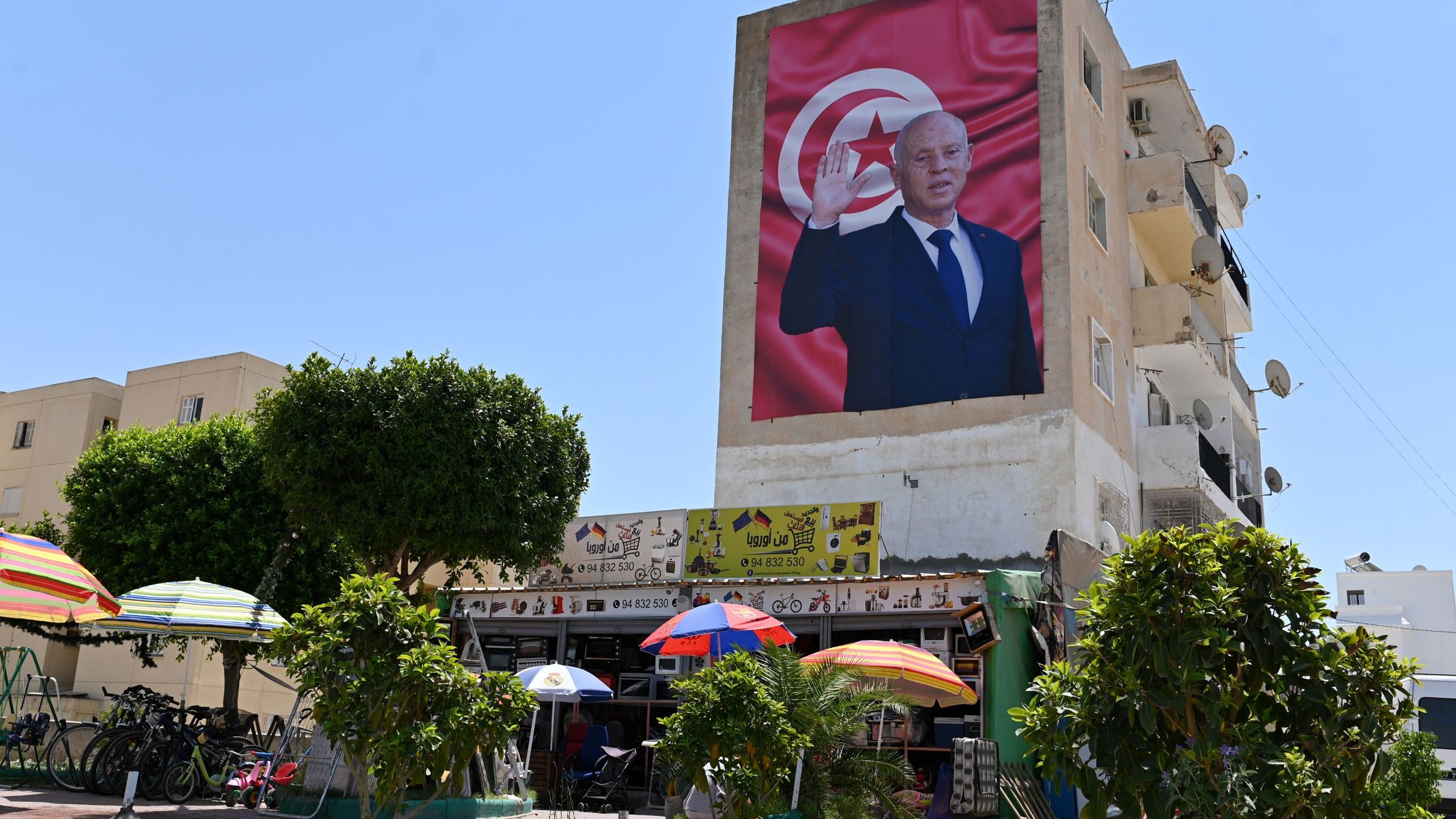 L'immagine mostra un grande murales su un edificio, raffigurante una figura maschile che saluta con una mano alzata. Sullo sfondo, è presente una bandiera che presenta i colori e il simbolo della Tunisia. Nella parte inferiore si possono vedere alcuni ombrelloni colorati, accanto a un'area con biciclette e persone, il tutto immerso in un ambiente urbano. Il cielo è sereno e luminoso, suggerendo una giornata di sole.