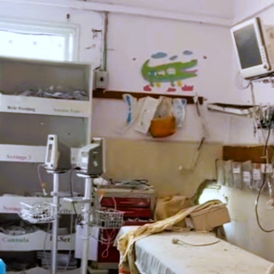 L'immagine mostra una stanza d'ospedale in uno stato di trascuratezza. Si possono notare letti ospedalieri con lenzuola disordinate, attrezzature mediche sparse e un mobile con vari oggetti all'interno. Le pareti sono decorate con disegni infantili, ma l'ambiente appare comunque piuttosto disordinato. Un monitor è appeso al muro e ci sono altri strumenti medici, indicando che si tratta di un'area destinata alla cura dei pazienti. La luce naturale entra dalla finestra, creando un contrasto con l'aspetto poco curato della stanza.