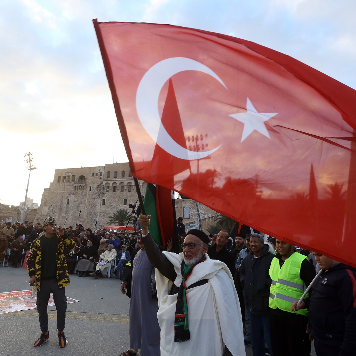 L'image montre une scène de rassemblement ou de manifestation en plein air. Au premier plan, un homme âgé tient fièrement un grand drapeau turc, qui est rouge avec une étoile et un croissant blancs. On peut apercevoir une foule de personnes en arrière-plan, certaines portant des vêtements de sécurité fluorescents. La scène se déroule peut-être dans un lieu historique, avec un bâtiment ancien visible derrière la foule. L'ambiance semble dynamique et engagée.