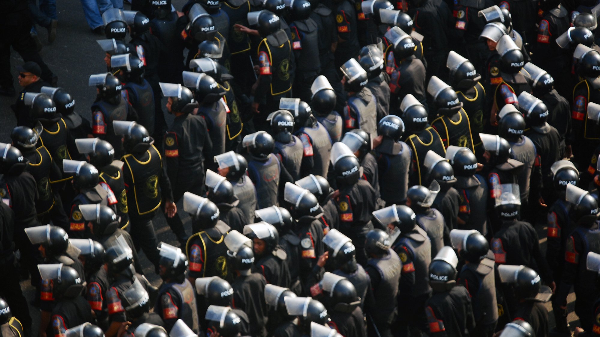 L'image montre un grand groupe de policiers en tenue d'émeute. Ils portent des casques et des boucliers, et sont regroupés de manière organisée. Les uniformes sont sombres, avec des détails visibles tels que des insignes ou des logos indiquant leur affiliation. L'ensemble donne une impression de préparation et de contrôle, évoquant un contexte de maintien de l'ordre lors d'une situation potentiellement tendue.