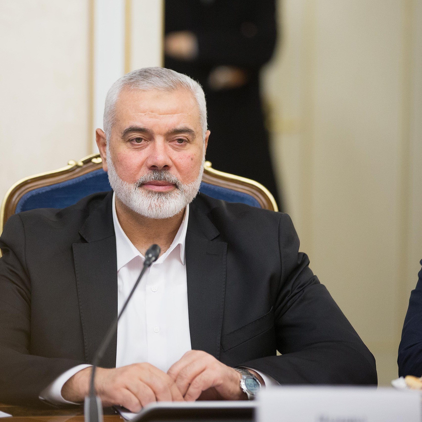 در تصویر، مردی با ریش خاکستری و موهای کوتاه دیده می‌شود که در حال نشستن در یک جلسه یا کنفرانس است. او ظاهری جدی دارد و یک کت تیره به تن دارد. بر روی میز مقابل او بطری‌های آب و تعدادی خوراکی دیده می‌شود. فضای اطراف به نظر رسمی و مرتبط با مذاکرات یا بحث‌های مهم می‌رسد.