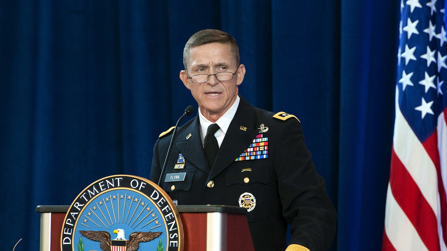 The image shows a man in a military uniform standing at a podium. The podium has the emblem of the Department of Defense prominently displayed. The man is speaking, possibly at a formal event or press conference. Behind him, there is a blue backdrop, and an American flag is visible to the side. The setting appears to be official and ceremonial.