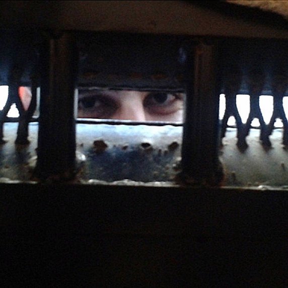 L'image montre un regard émergeant à travers une grille ou une ouverture sombre. Les yeux de la personne sont bien visibles, tandis que le reste de son visage est caché par l'ombre ou la structure. Les bords de l'ouverture semblent usés, ajoutant une atmosphère de confinement ou de mystère à la scène.