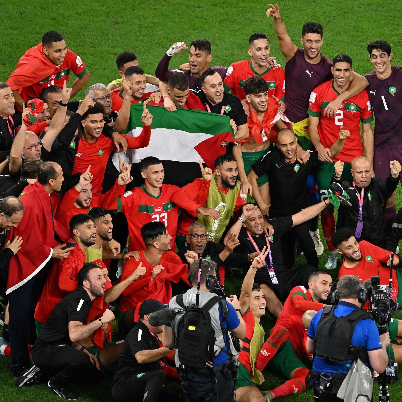 تظهر في الصورة مجموعة من اللاعبين والمشجعين المغاربة يحتفلون بشكل jubilant. يحمل اللاعبون العلم المغربي ويظهر عليهم الفرح والانتصار. يتمركز بعضهم حول مدربهم أو مدربين، بينما يتواجد آخرون بالقرب من كاميرات التصوير، مما يوحي بأجواء احتفالية قوية بعد تحقيق إنجاز رياضي. تعكس الصورة روح الفريق والوحدة بين اللاعبين.
