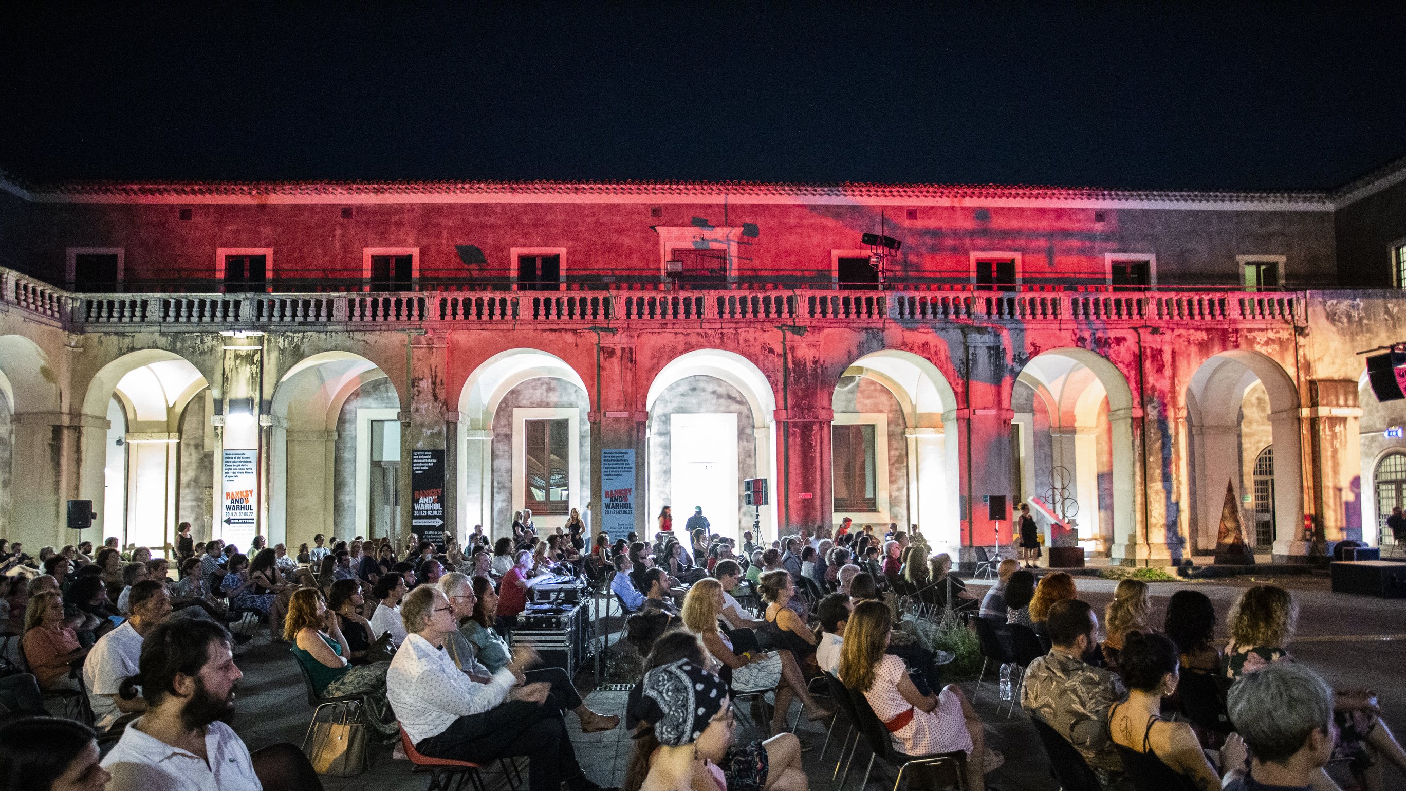 L'immagine mostra una grande sala all'aperto, con archi eleganti che circondano lo spazio. È sera e l'ambiente è illuminato da luci soffuse, creando un'atmosfera suggestiva. In primo piano, un folto pubblico è seduto su sedie disposte in file, mentre alcuni spettatori osservano attentamente. La scena trasmette un senso di partecipazione a un evento culturale o a una performance artistica. I dettagli architettonici dell'ambiente circostante sono ben visibili, contribuendo all'eleganza della location.