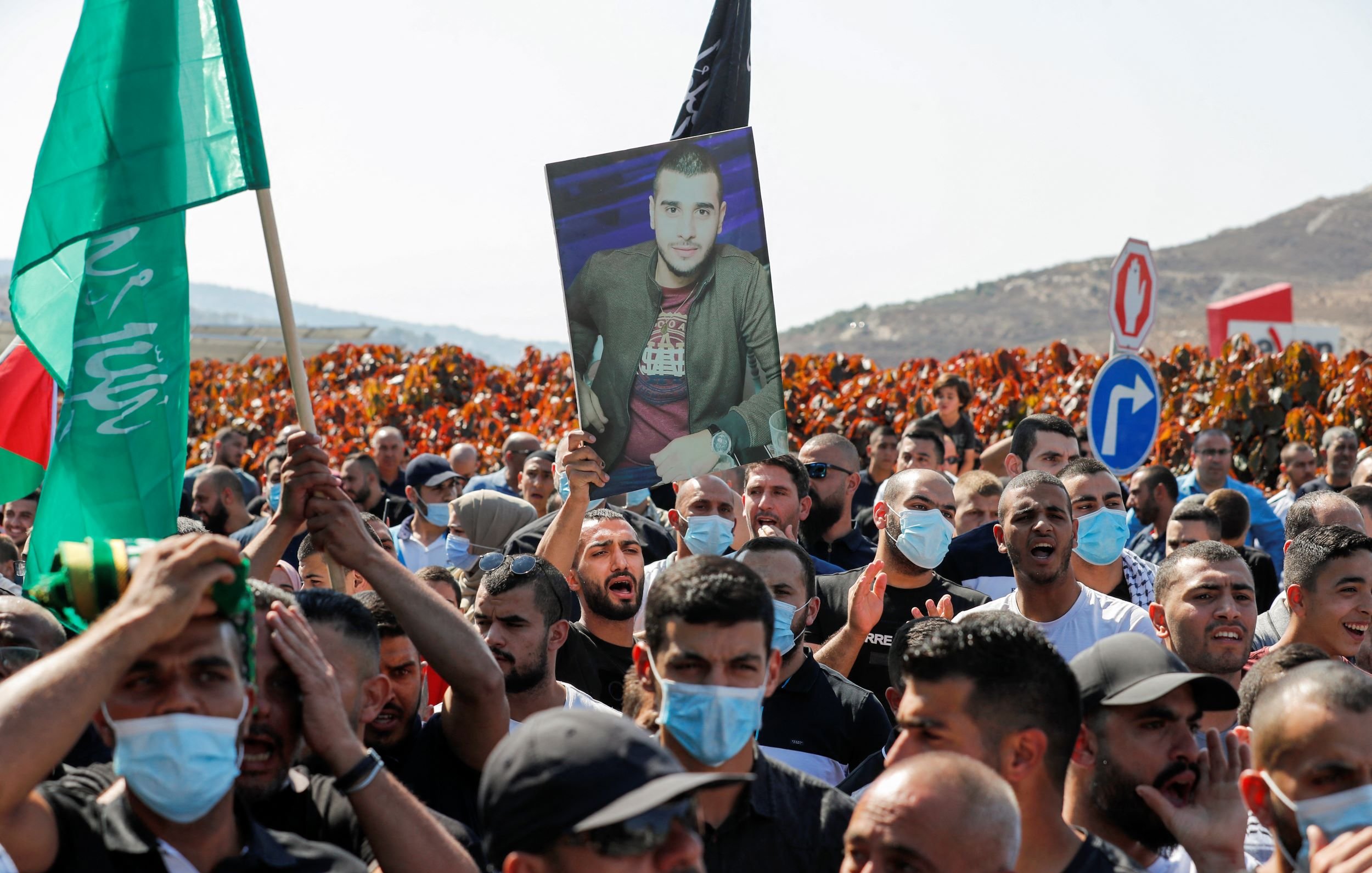 در تصویر، گروهی از افراد در یک تظاهرات حضور دارند. تعداد زیادی از افراد پرچم‌هایی به رنگ‌های مختلف در دست دارند. یکی از تظاهرکنندگان تصویری از یک مرد را بالا نگه داشته است. بیشتر افراد ماسک به صورت دارند و در پس‌زمینه، زمین‌های سرسبز و تپه‌هایی دیده می‌شود. به نظر می‌رسد که این تجمع به منظور اعتراض یا یادبود برگزار شده باشد.