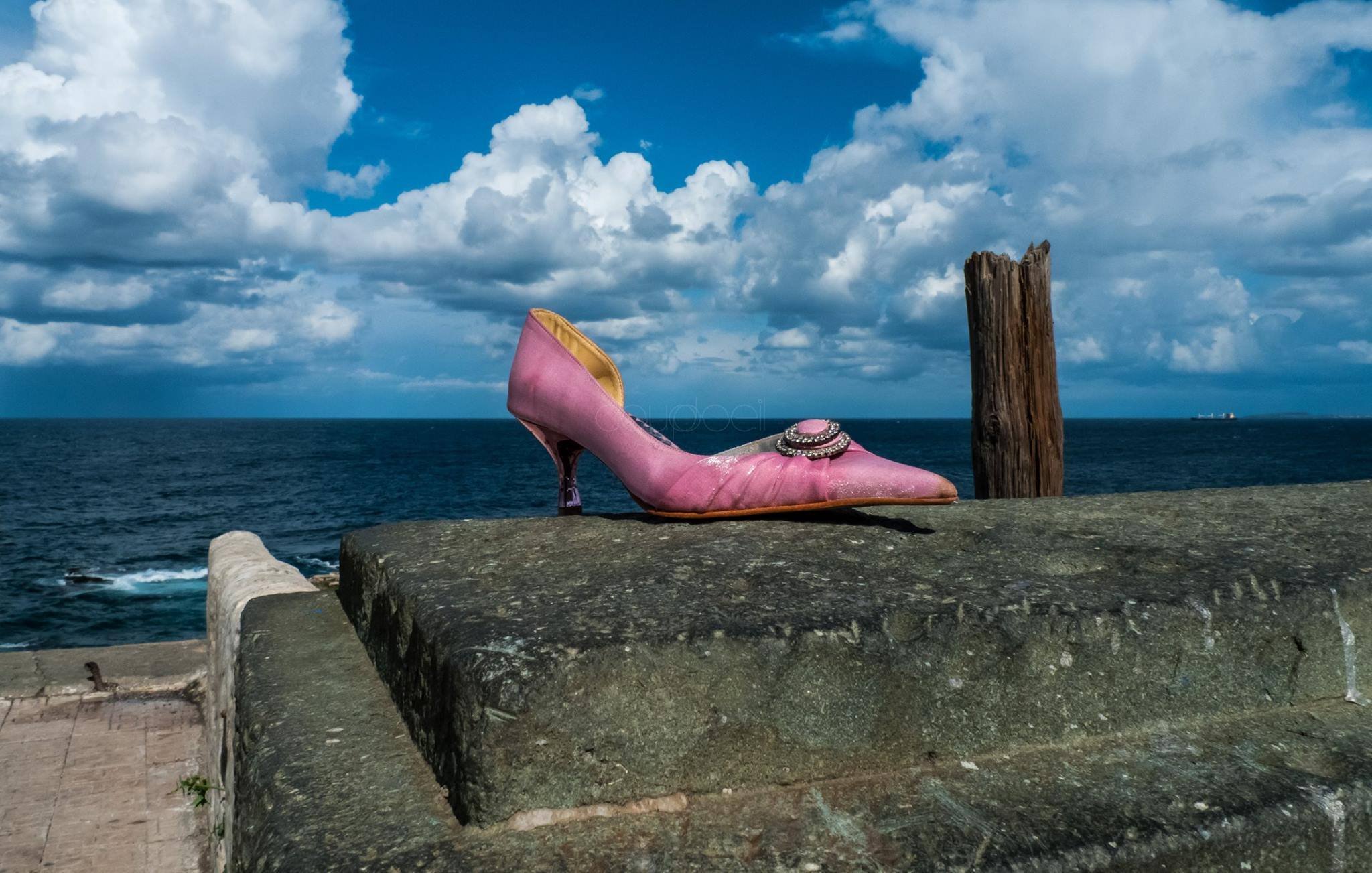 L'image montre une chaussure à talon rose, posée sur un mur en pierre. En arrière-plan, on peut voir une mer calme sous un ciel rempli de nuages blancs et d'un bleu éclatant. À droite, un poteau en bois émerge de la surface, ajoutant une touche de rusticité au paysage. L'ensemble évoque un mélange de solitude et de beauté naturelle.