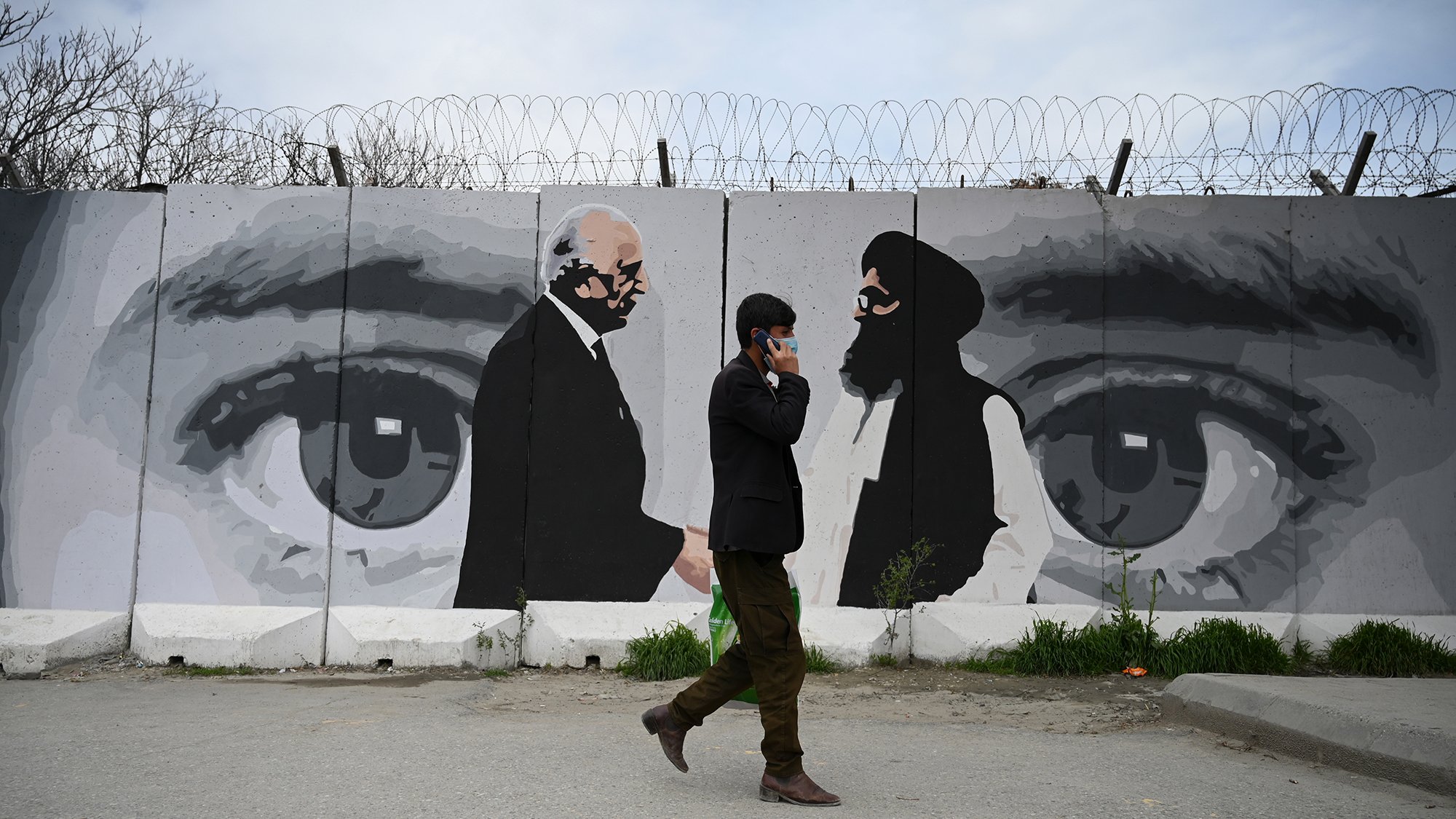 تظهر في الصورة حائطًا مزخرفًا برسوم جدارية لشخصيتين تتبادلان النظرات، حيث توجد عيون كبيرة في الخلفية تعكس مشاعر متباينة. في المقدمة، يمشي رجل يرتدي بدلة سوداء، ممسكًا بهاتفه المحمول. الحائط يتضمن أسلاك شائكة، مما يضيف إحساسًا بالانقسام أو التوتر. تبرز الألوان الرمادية والأبيض والأسود بشكل كبير، مما يضفي طابعًا دراماتيكيًا على المشهد.