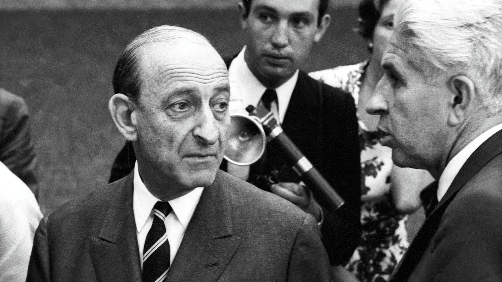 Deux hommes en costume, discutant sérieusement, entourés d'autres personnes. Époque rétro.