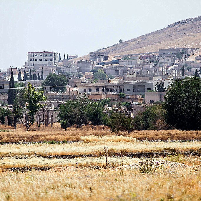 L'image montre un paysage rural avec des bâtiments en arrière-plan, probablement une petite ville ou un village. Au premier plan, il y a des champs avec de l'herbe sèche ou des cultures en attente de récolte. On peut voir une colline à l'arrière-plan, ainsi qu'un réservoir d'eau. La végétation est présente, avec quelques arbres éparpillés. L'atmosphère semble calme et ensoleillée, dégageant une ambiance de milieu rural.