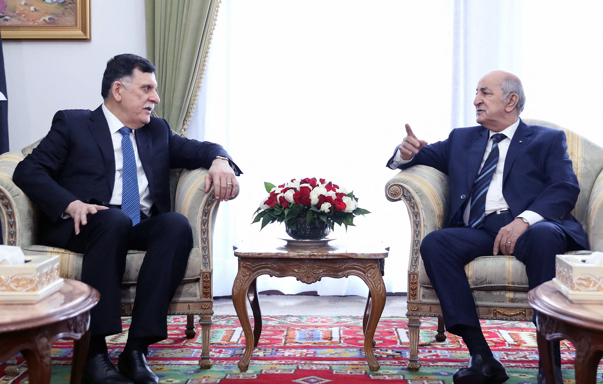 L'image montre deux hommes assis dans un environnement décoré de façon formelle. Ils sont en train de discuter, chacun semblant attentif à l'autre. Le premier homme, habillé en costume avec une cravate, se tourne légèrement vers le second. Ce dernier, également en costume, gesticule avec une main, ce qui pourrait indiquer qu'il est en train de faire un point ou de partager une idée. Entre eux, il y a une table basse ornée d'un arrangement floral. L'arrière-plan est lumineux et dégage une ambiance de rencontre officielle. Des éléments décoratifs, comme des rideaux et un tapis, ajoutent une touche élégante à la scène.