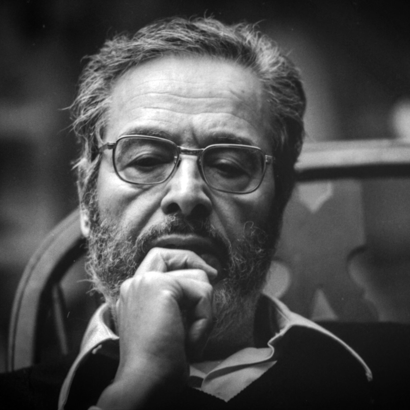 The image features a man with a thoughtful expression, sitting in a wooden chair. He has a beard and glasses, and he appears to be deep in contemplation. The lighting is soft, creating a moody atmosphere, and the background is blurred, focusing attention on his contemplative demeanor. The photograph has a classic black-and-white aesthetic, emphasizing the seriousness of the moment.