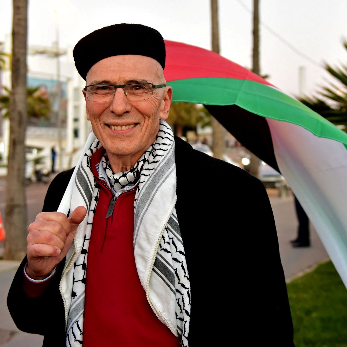 Un homme souriant porte un keffieh, derrière lui un drapeau palestinien sur fond urbain.