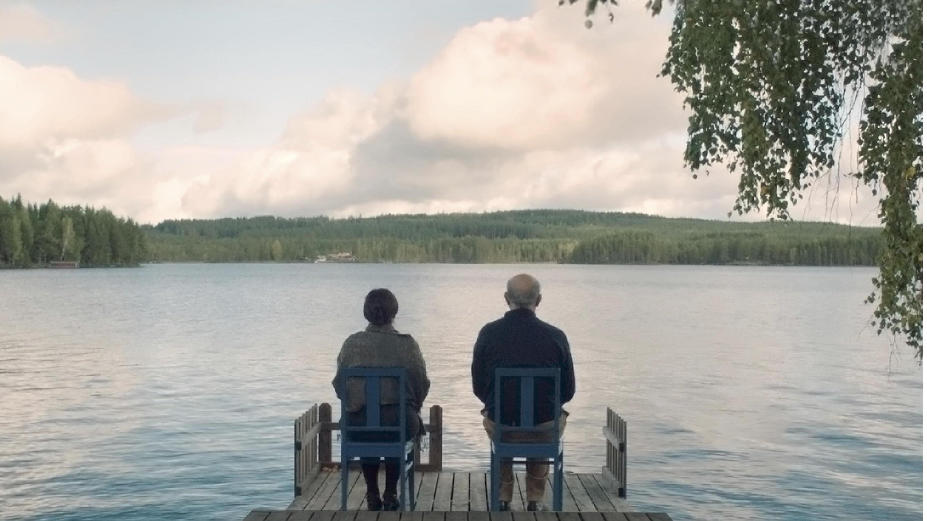 L'image montre deux personnes assises sur des chaises en bois sur une jetée qui s'avance dans un lac paisible. Ils regardent vers l'horizon, avec des arbres verdoyants en arrière-plan et un ciel nuageux au-dessus. L'atmosphère est calme et sereine, évoquant un moment de contemplation ou de discussion tranquille.