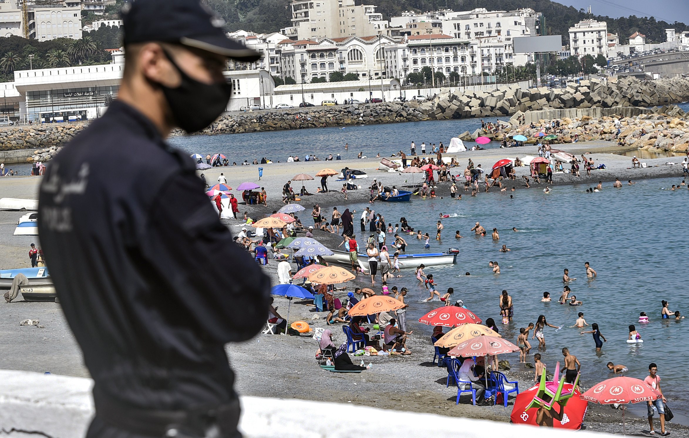 L'image montre un agent de sécurité en premier plan, portant un masque, surveillant une plage animée. En arrière-plan, des familles et des groupes de personnes profitent du temps ensoleillé, se baignant dans l'eau et se reposant sous des parasols colorés. On aperçoit également des bateaux au loin et des bâtiments sur la côte qui ajoutent à l'ambiance estivale de la scène.