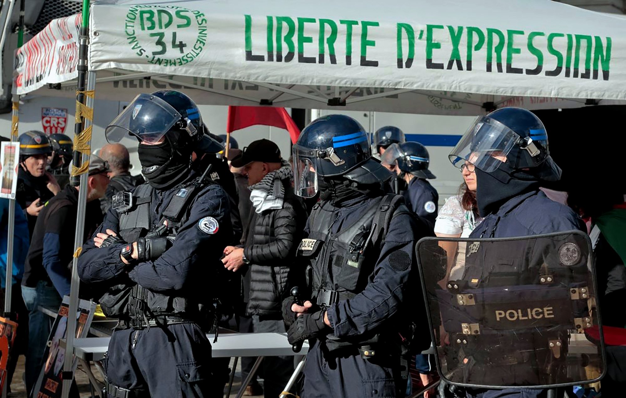 L'image montre un groupe de policiers en uniforme, équipés de casques et de protections, se tenant devant un stand ou une tente avec l'inscription "LIBERTE D'EXPRESSION". On peut voir plusieurs personnes en arrière-plan, probablement des manifestants ou des observateurs, dans un environnement urbain. L'atmosphère semble être celle d'un rassemblement ou d'une manifestation, avec des thèmes liés à la liberté d'expression.
