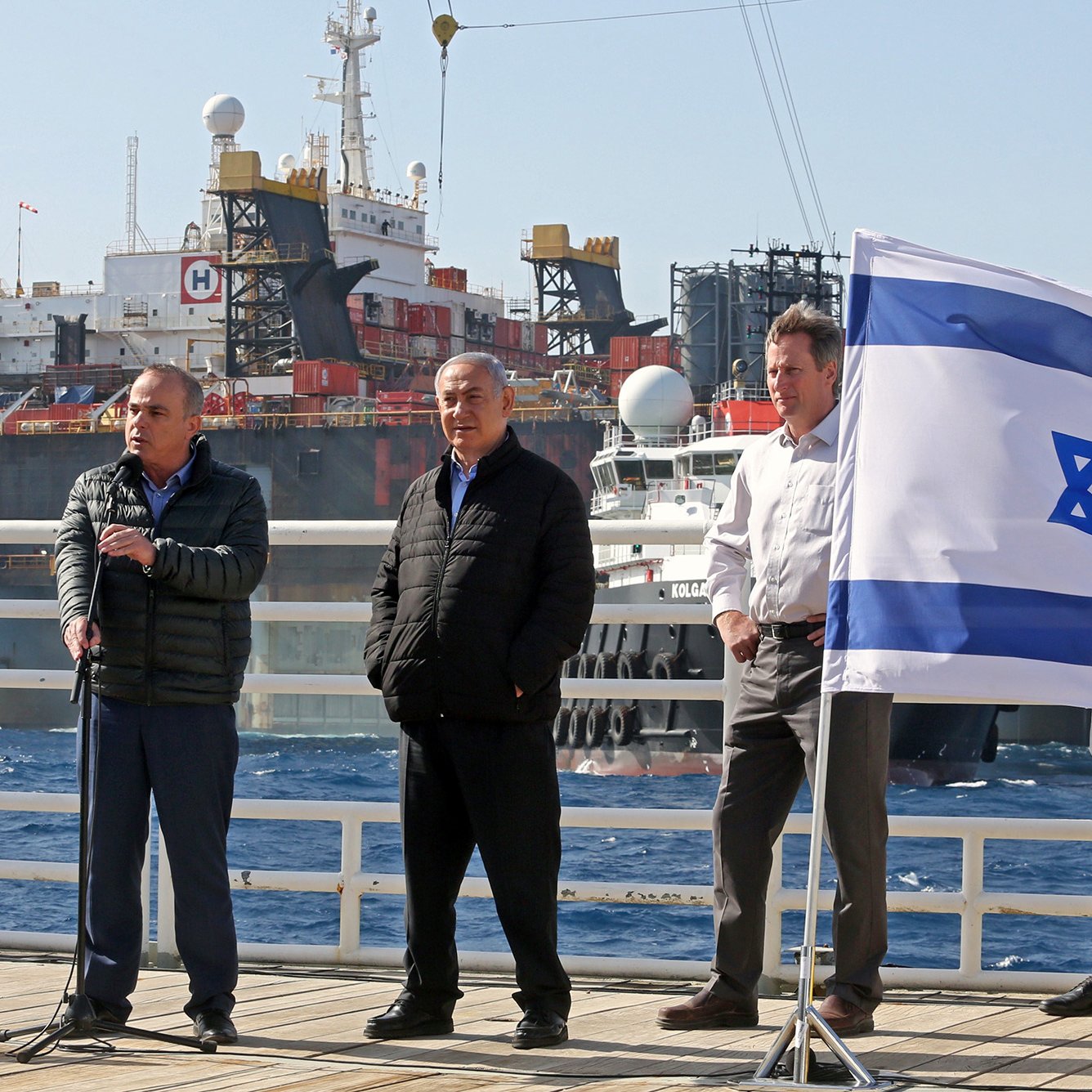 L'image montre plusieurs personnes sur un quai, en face de navires en arrière-plan. À gauche, un homme s'exprime avec un micro, tandis qu'un autre homme se tient à ses côtés, visiblement en position d'écoute. Un troisième homme est également présent. Au premier plan, un drapeau israélien flotte, ajoutant une touche patriotique à la scène. Les navires derrière eux semblent être engagés dans des opérations maritimes, suggérant un contexte lié à l'industrie maritime ou énergétique. L'ambiance paraît formelle, indiquant probablement un événement officiel ou une annonce importante.