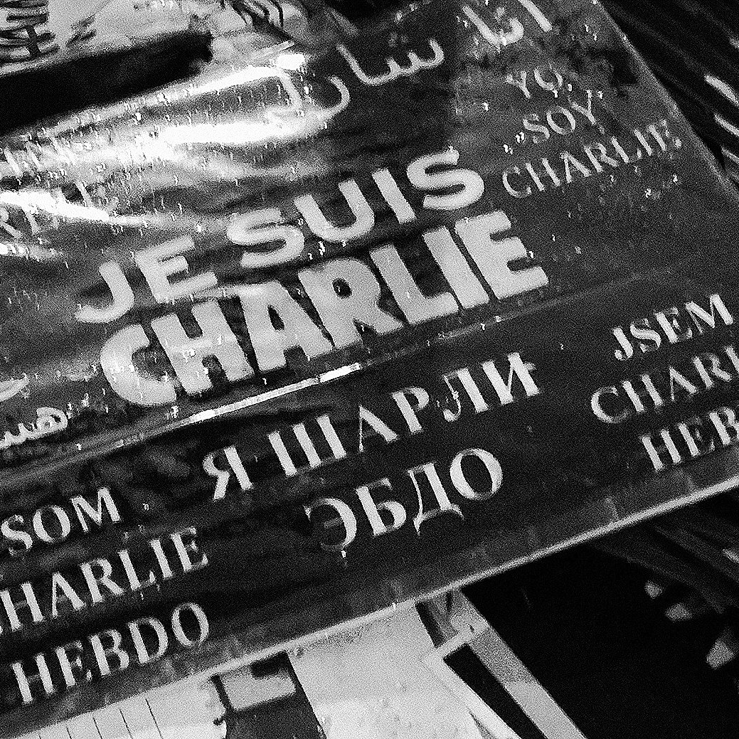 L'image montre une affiche ou un panneau avec l'inscription "JE SUIS CHARLIE" en plusieurs langues. Les mots sont écrits en noir sur fond blanc, et le tout a un aspect qui évoque le soutien et la solidarité, probablement en rapport avec les événements tragiques auxquels cette phrase fait allusion. On peut également voir des éléments environnants qui semblent être des morceaux de papier ou des affiches, ce qui donne un aspect de rassemblement ou de manifestation.