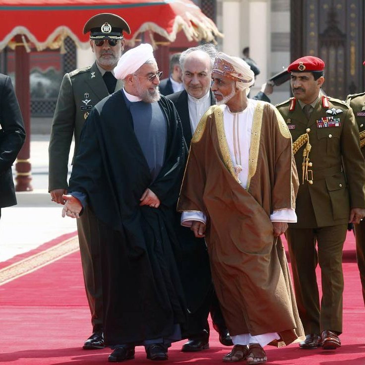 L'image montre un groupe de personnes marchant sur un tapis rouge. Au centre, deux hommes en discussion, l'un portant une robe noire et l'autre une robe traditionnelle d'Oman. Ils sont entourés de plusieurs membres de la sécurité et d'officiers en uniforme. Le cadre est formel, avec un décor architectural en arrière-plan qui suggère un événement diplomatique ou officiel.