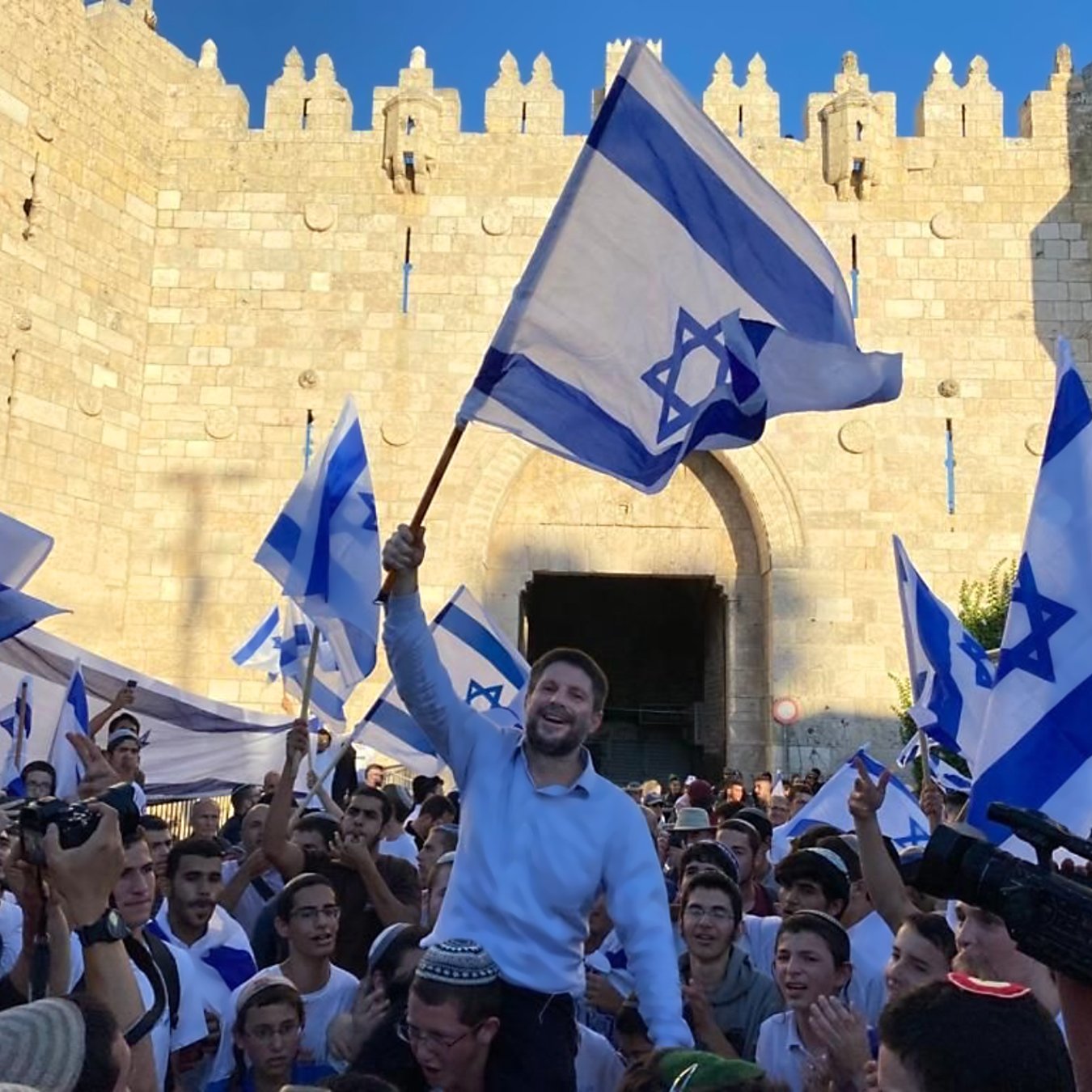 L'image montre une foule rassemblée devant une grande porte en pierre, probablement située dans une ville historique. Les participants tiennent des drapeaux bleu et blanc, représentant l'État d'Israël. Un homme se tient au centre, soulevant un drapeau avec un sourire, tandis que d'autres l'entourent, festoyant. L'atmosphère semble festive et joyeuse, suggérant une célébration ou un événement important.