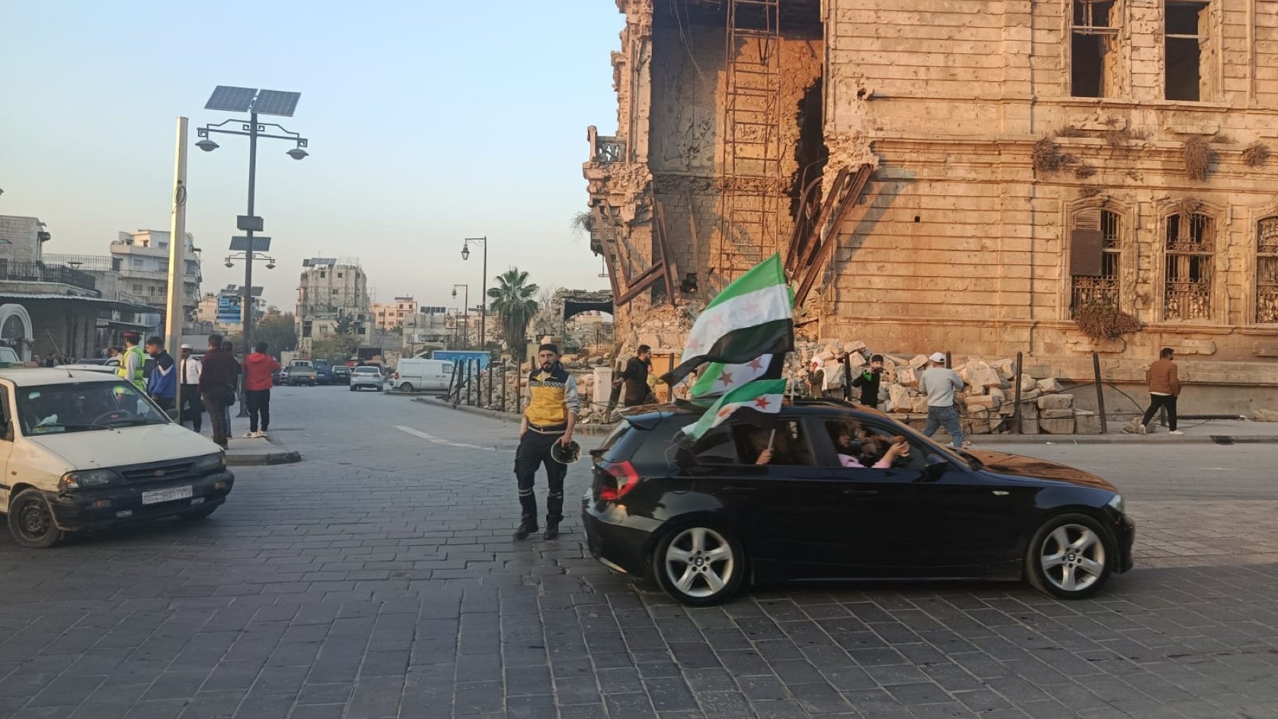Scène de rue avec un bâtiment endommagé, des gens et une voiture avec un drapeau.
