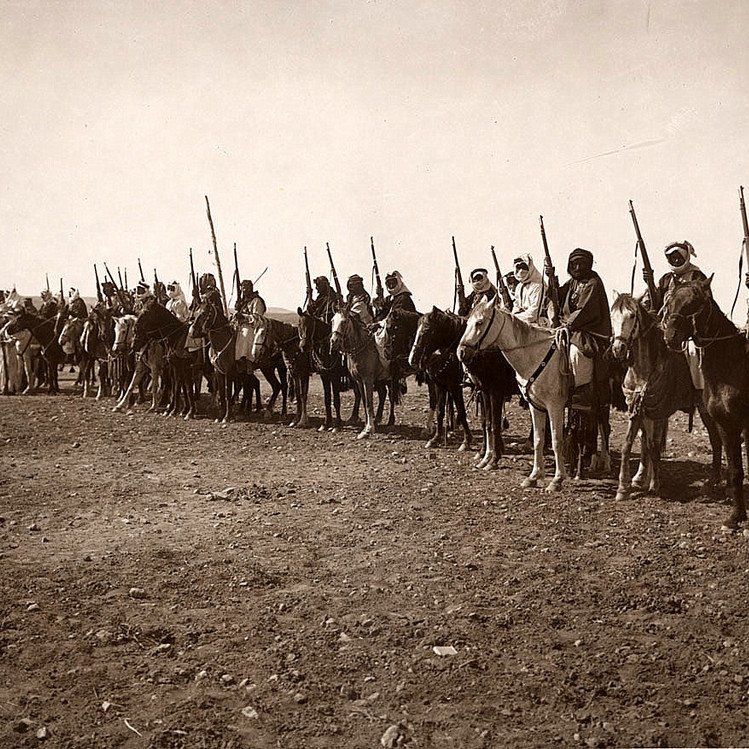 L'image montre une longue ligne de cavaliers en rang, tous montés sur des chevaux. Chaque cavalier est équipé d'une lance ou d'un bâton, et beaucoup portent des vêtements traditionnels. L'arrière-plan semble désertique, avec un sol rugueux et peu de végétation. L'atmosphère de l'image évoque une scène historique ou militaire, mettant en avant des guerriers ou des membres d'une tribu. Les cavaliers adoptent une posture fière et attentive, prêts pour une action ou une procession.