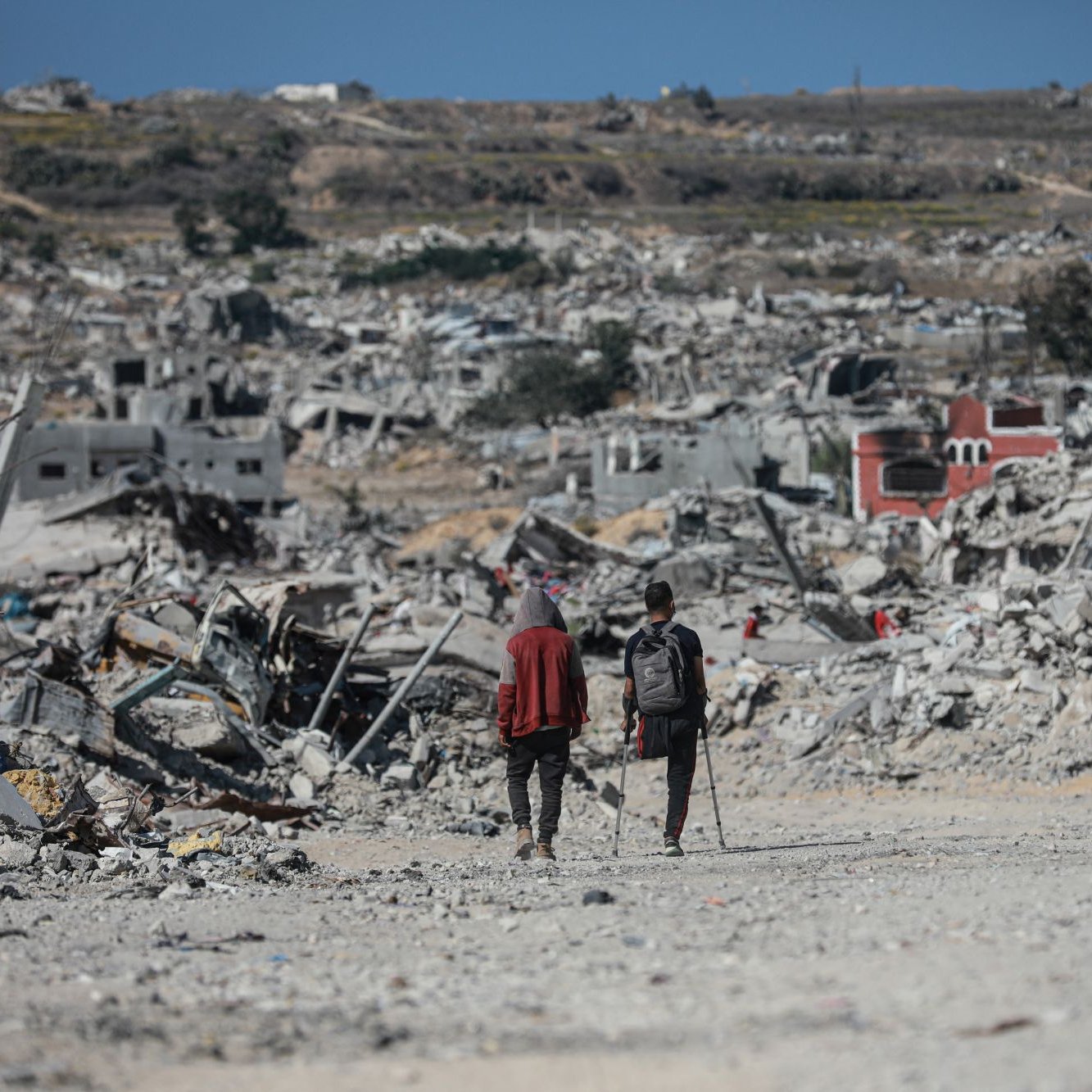 Two people walk through a vast landscape of destruction and rubble, with collapsed buildings around them.