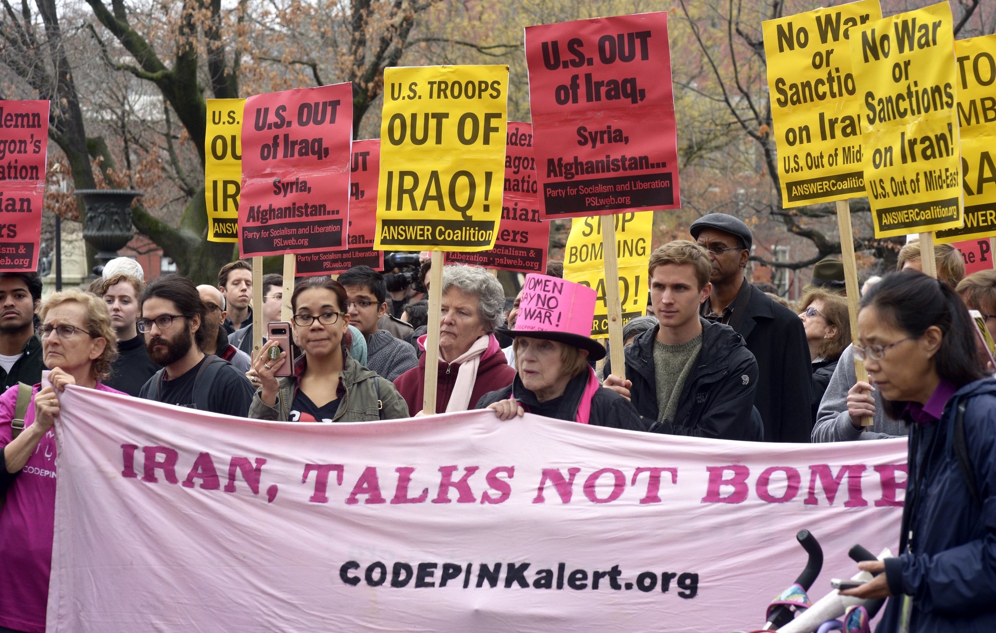 L'image montre une manifestation où des personnes tiennent des pancartes avec des slogans anti-guerre. Plusieurs affiches demandent le retrait des troupes des États-Unis d'Irak, de Syrie et d'Afghanistan. Les manifestants semblent déterminés et unis dans leur message, qui appelle à la paix, avec un grand banner rose affichant "IRAN, TALKS NOT BOMBS" et le site web de CODEPINK. L'atmosphère semble à la fois sérieuse et engagée.