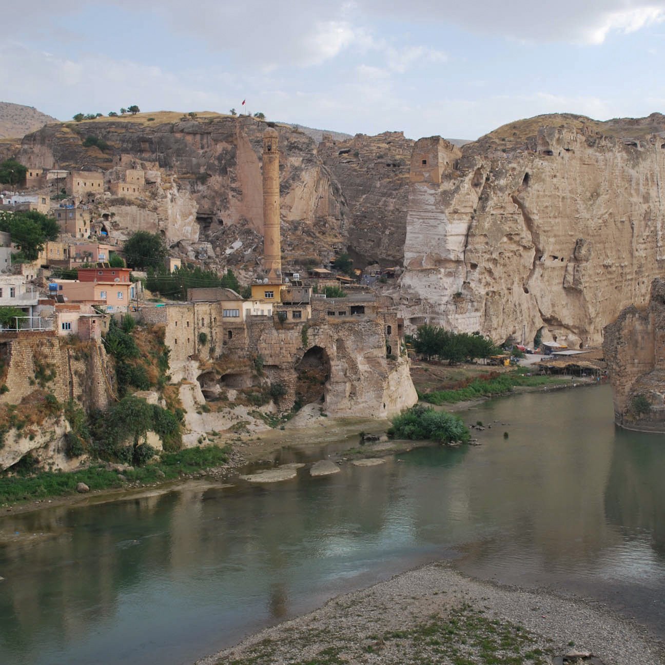 L'image montre un paysage pittoresque avec des formations rocheuses impressionnantes en arrière-plan. On y voit un village construit sur les flancs de la montagne, avec des maisons aux couleurs chaudes. Un fleuve calme coule au premier plan, reflétant les alentours. Quelques arbres verdoyants et des ruines anciennes ajoutent à la beauté historique de la scène. L'atmosphère est tranquille, dominée par les teintes naturelles des roches et de l'eau.