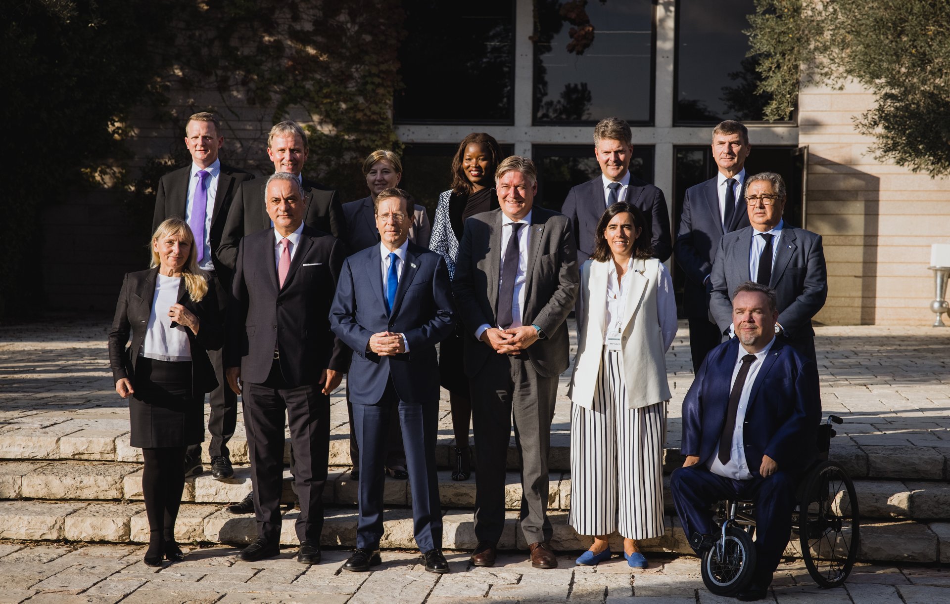 L'image montre un groupe de personnes posant ensemble à l'extérieur, sur des marches devant un bâtiment. Il y a dix-sept individus, dont certains portent des costumes et d'autres des tenues variées. L'arrière-plan est un jardin ou un espace extérieur avec des arbres et une architecture moderne. Les personnes affichent des expressions souriantes, ce qui suggère un moment de camaraderie ou une occasion formelle, comme une réunion ou un sommet.