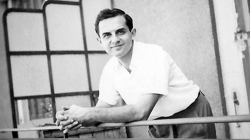 L'image montre un jeune homme posant avec un sourire amical. Il est vêtu d'une chemise blanche et de pantalons clairs, se tenant sur un balcon avec des barres métalliques. L'arrière-plan présente une architecture simple, avec des fenêtres et des volets. L'ambiance est décontractée et ensoleillée, suggérant un moment de détente.