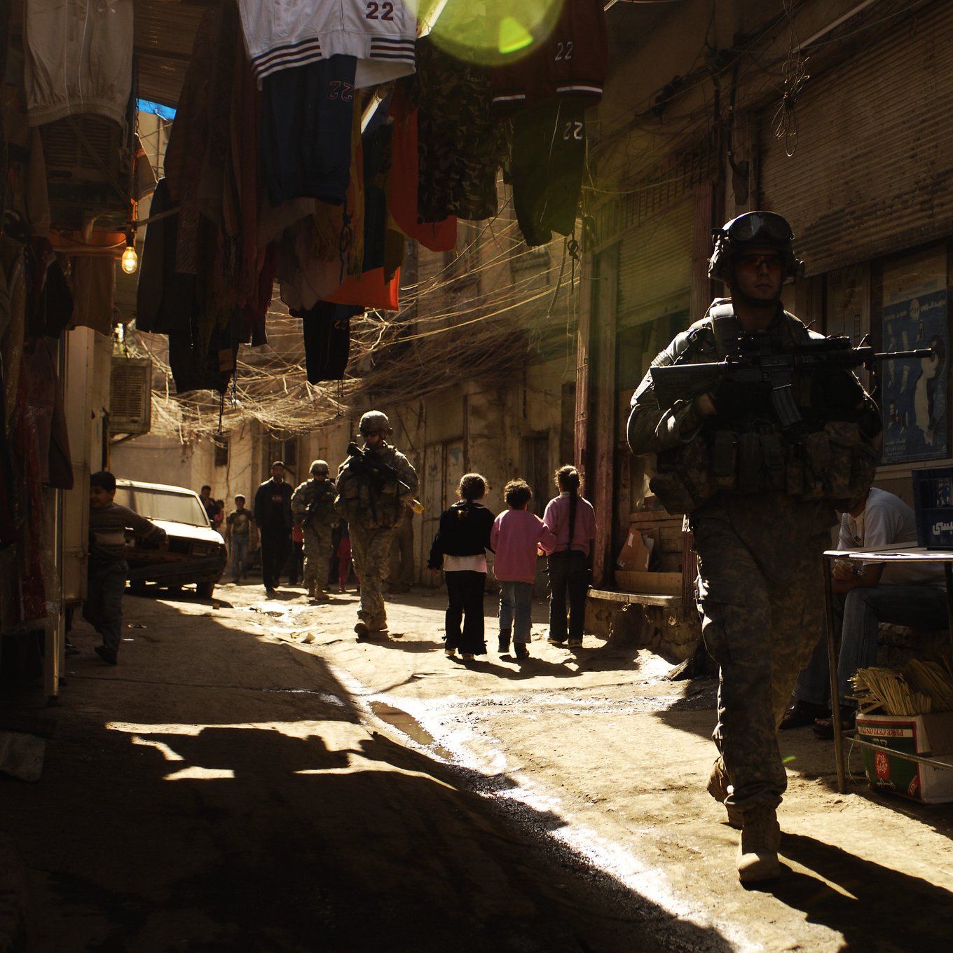 تظهر الصورة مشهداً في حي شعبي، حيث يسير جنود يرتدون زيًا عسكريًا ويحملون أسلحة. تضيء أشعة الشمس المساحة بين المباني، مما يخلق تأثيرًا جميلًا من الضوء والظل. في الخلفية، يمكن رؤية مجموعة من الأشخاص، يبدو أن بعضهم أطفال، مما يوحي بحياة يومية في هذا المكان. كما تظهر الملابس المعلقة على الجانبين، مما يعطي انطباعًا عن الحياة المحلية والتجارة في هذا الحي.