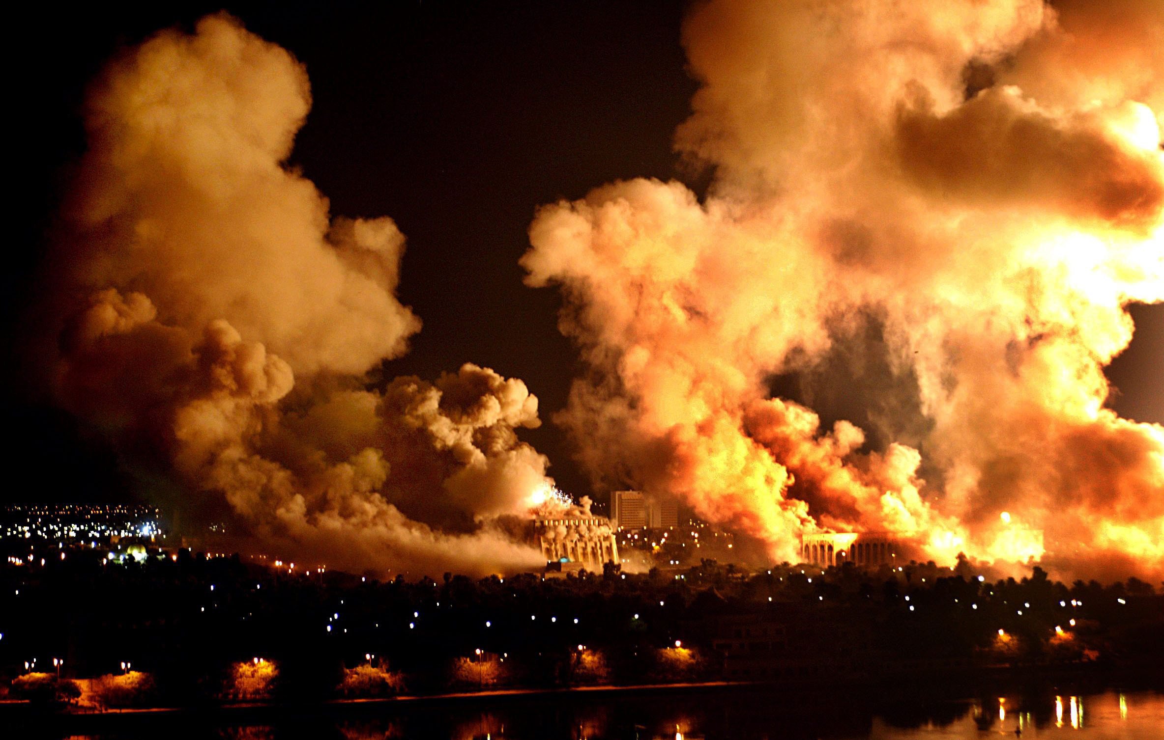 L'image montre une scène nocturne marquée par d'épais nuages de fumée et des flammes. On peut apercevoir des explosions ou des incendies créant une lumière intense dans l'obscurité. Les perturbations dans le ciel semblent témoigner d'un événement violent, tandis qu'en bas, on distingue une ville avec des lumières qui contrastent avec le chaos. L'ensemble dégage une atmosphère dramatique et inquiétante.