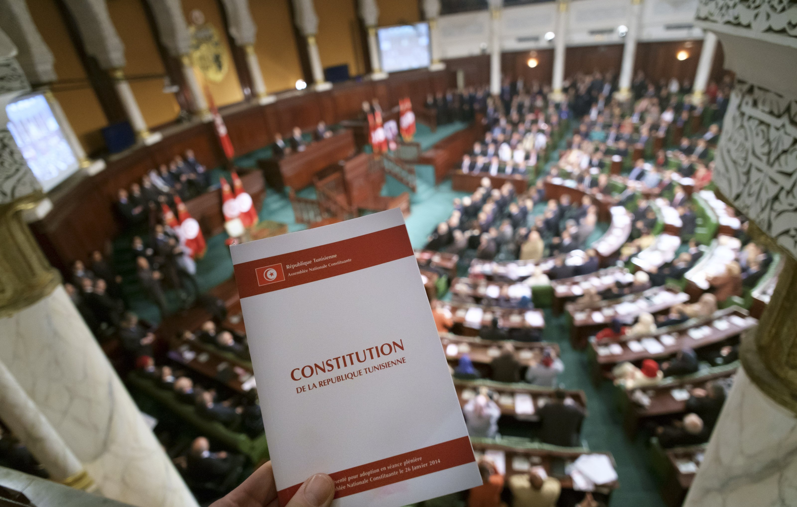 تظهر في الصورة قاعة البرلمان التونسي، حيث يوجد مجموعة كبيرة من النواب والموظفين في الجلسة. في المقدمة، هناك شخص يحمل نسخة من الدستور التونسي. الخلفية مليئة بالعلم التونسي، مما يدل على أهمية الحدث الذي يتم مناقشته في القاعة. التصميم الداخلي للقاعة يبدو أنيقًا، مع تفاصيل معمارية تقليدية.